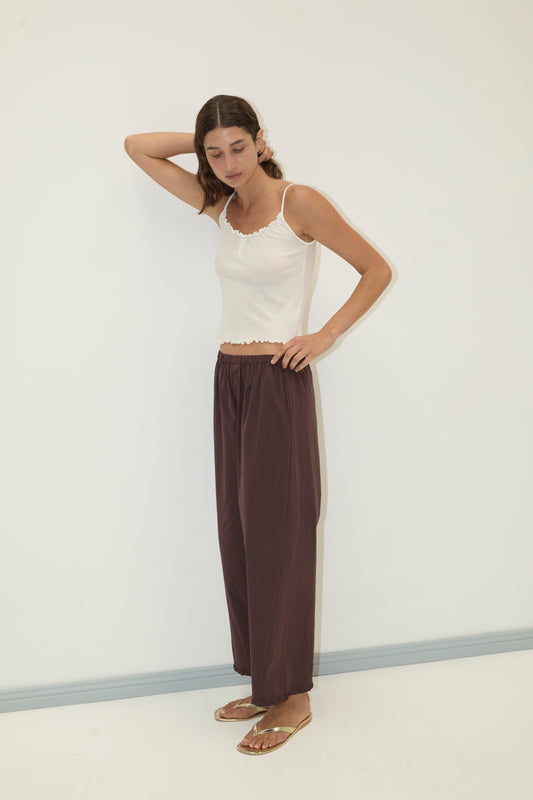 A woman stands against a plain white wall, wearing the Flounce Tank - Cream by Deiji Studios, loose brown pants, and gold sandals. She has one hand behind her head and looks downward with a relaxed expression.