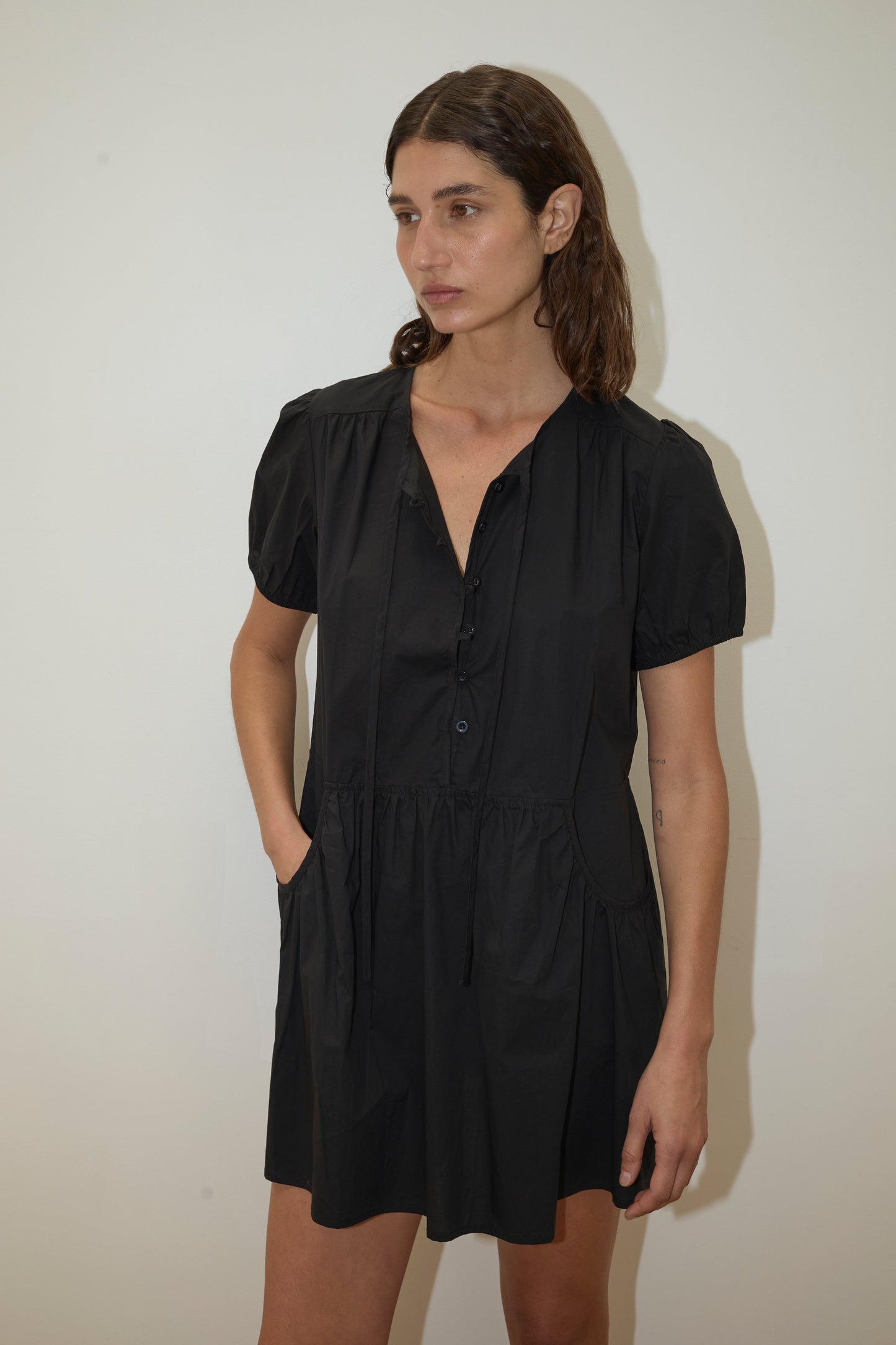 A woman with brown hair stands against a white background, wearing Deiji Studios' Pocket Mini Dress - Black with short sleeves, a gathered waist, side pockets, and made from organic cotton. Her left hand is in her pocket and her expression is neutral.