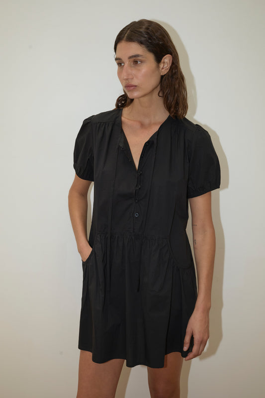 A woman with brown hair stands against a white background, wearing Deiji Studios' Pocket Mini Dress - Black with short sleeves, a gathered waist, side pockets, and made from organic cotton. Her left hand is in her pocket and her expression is neutral.