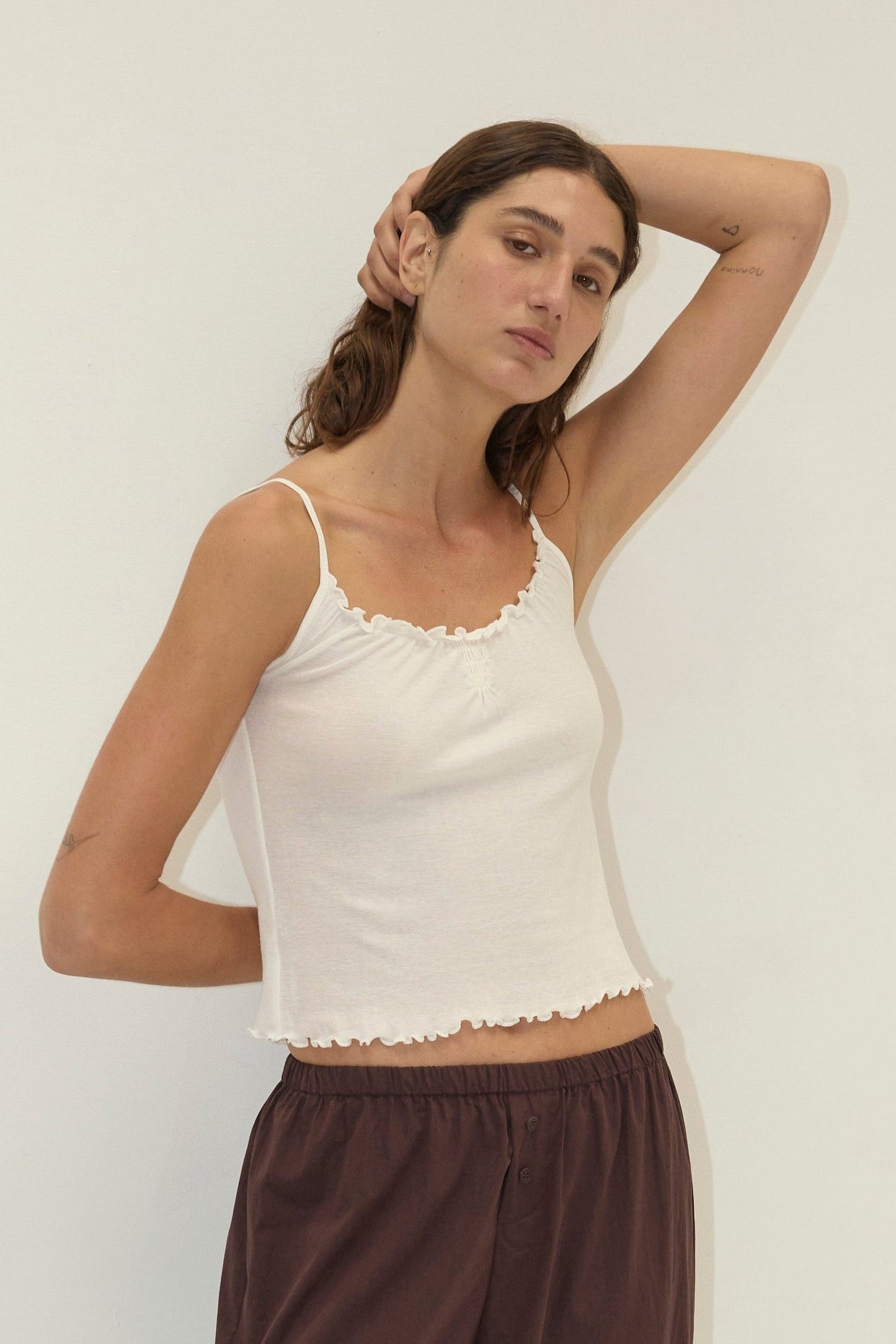 A woman with long brown hair poses against a plain white wall, wearing the Flounce Tank - Cream by Deiji Studios and loose maroon pants. One hand rests behind her head, and she has a neutral expression.