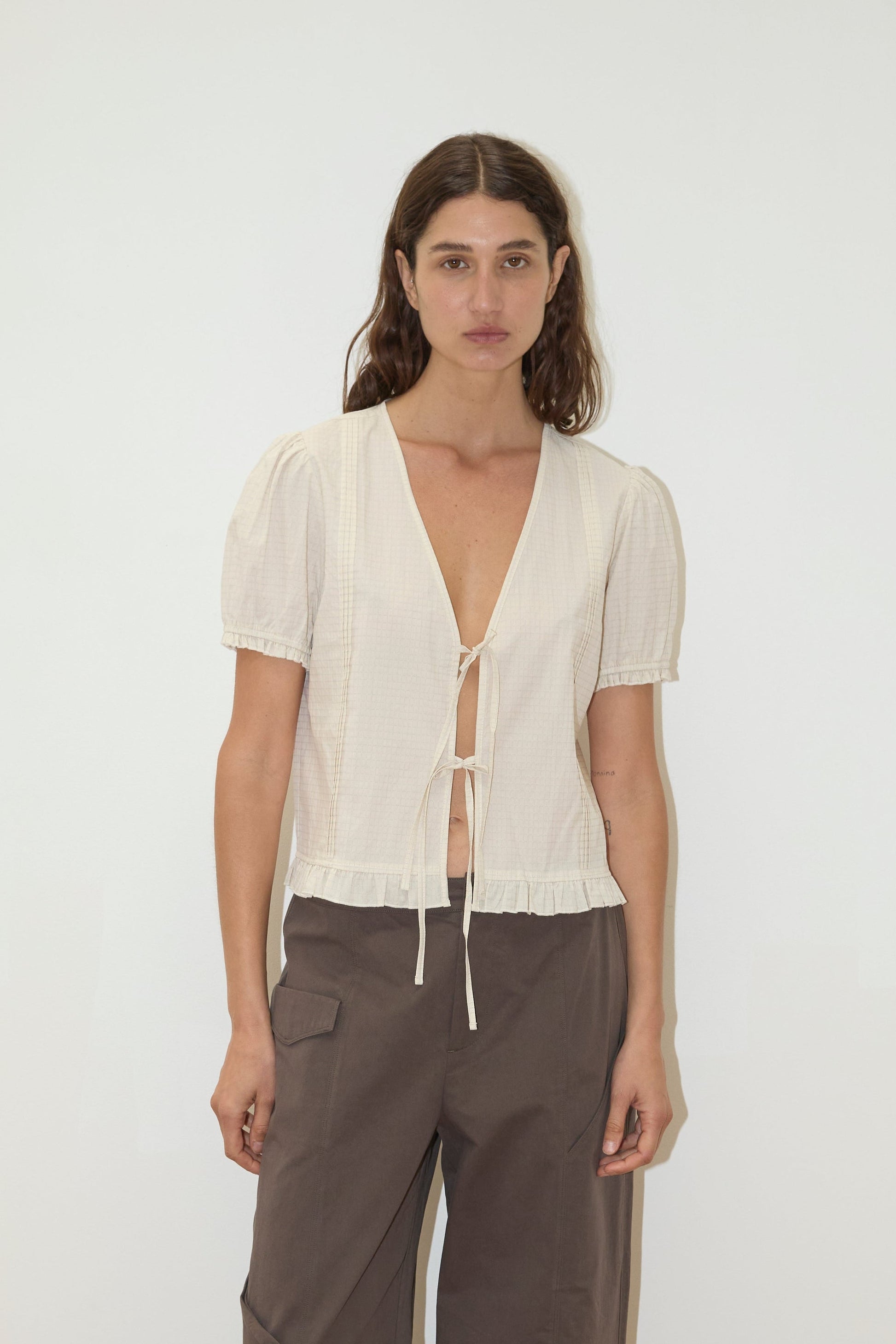 A woman stands against a plain white background, wearing the Pintuck Top - Bone Check by Deiji Studios, a light beige short-sleeve V-neck pintuck top with a tie front and ruffled hem, paired with loose-fitting brown pants.