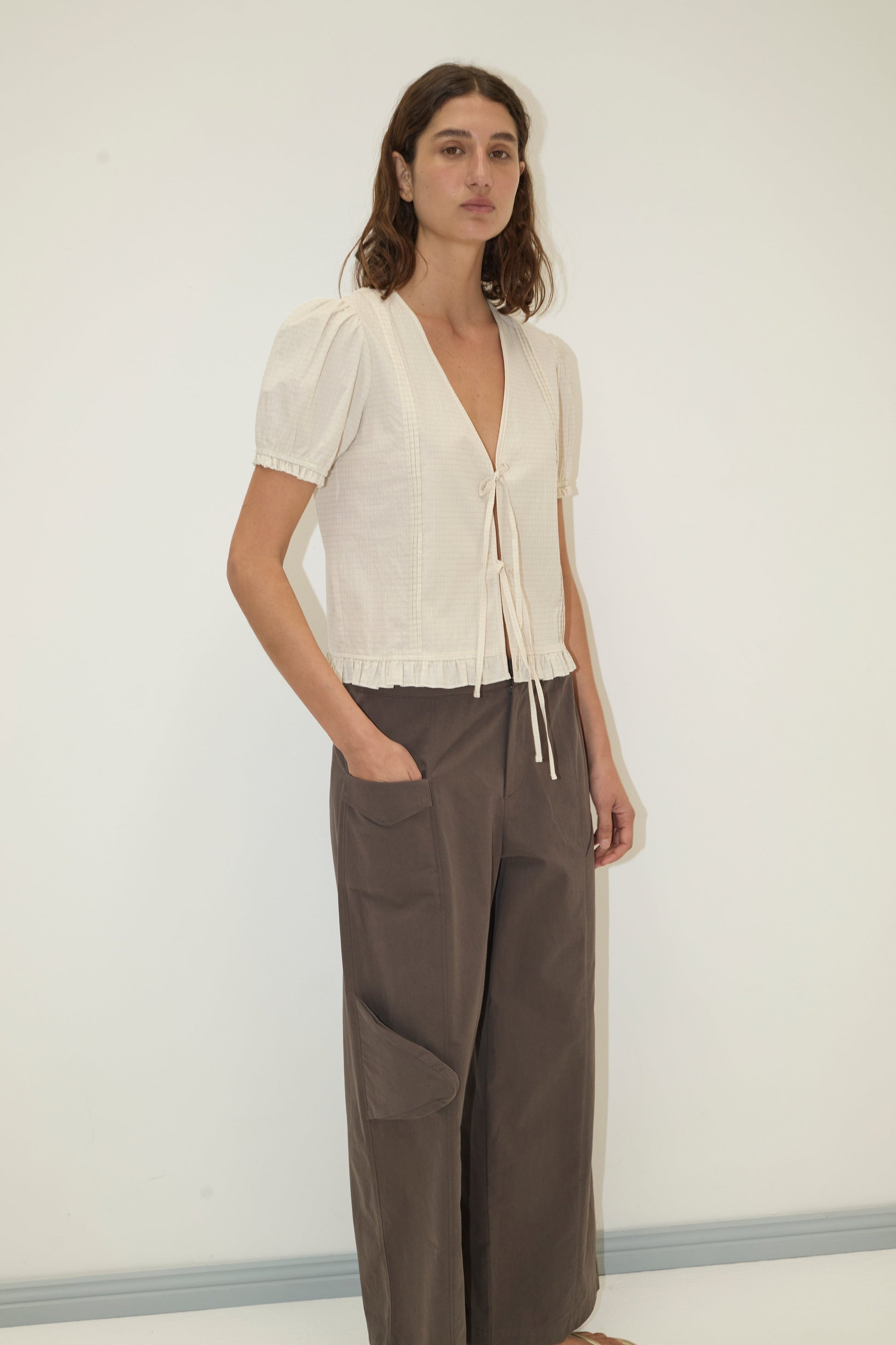 A woman stands against a white wall, wearing the Deiji Studios Pintuck Top - Bone Check in organic cotton with a front tie and loose brown wide-leg pants. She has long, wavy brown hair and one hand in her pocket.