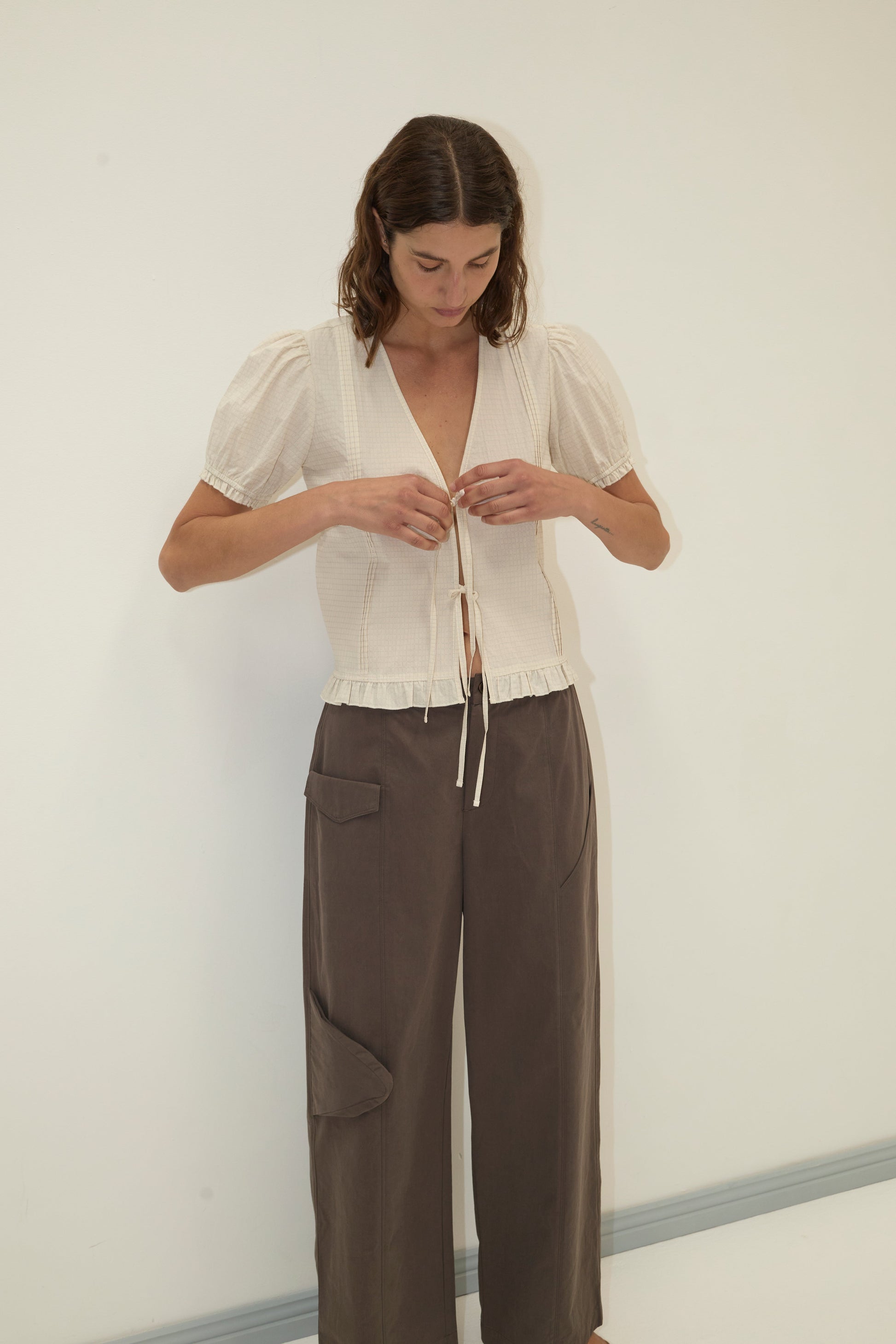 A woman stands against a plain light wall, looking down as she ties the front of her Pintuck Top - Bone Check. She pairs the top with high-waisted, wide-leg brown pants featuring large pockets.