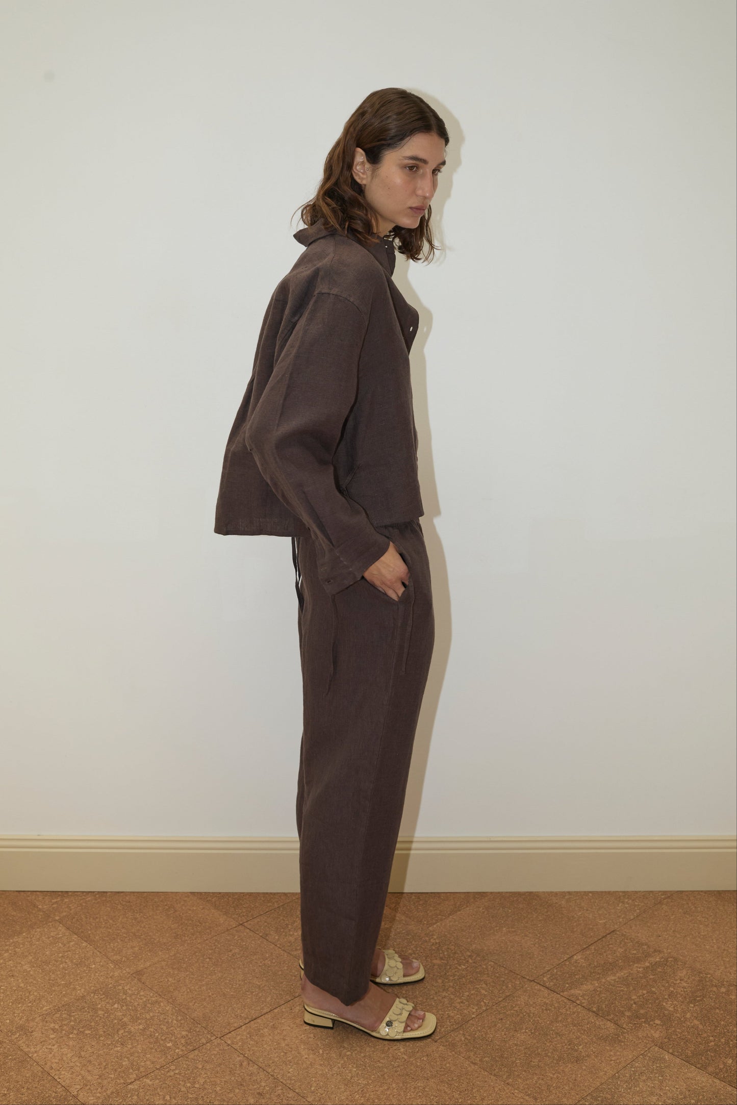 A person with wavy brown hair stands sideways against a plain white wall, wearing the Deiji Studios 4 Tie Set - Bark, a loose organic linen set in dark brown, paired with beige low-heeled sandals and hands in pockets.