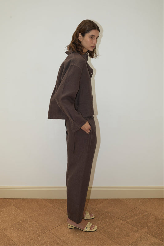 A person with wavy brown hair stands sideways against a plain white wall, wearing the Deiji Studios 4 Tie Set - Bark, a loose organic linen set in dark brown, paired with beige low-heeled sandals and hands in pockets.