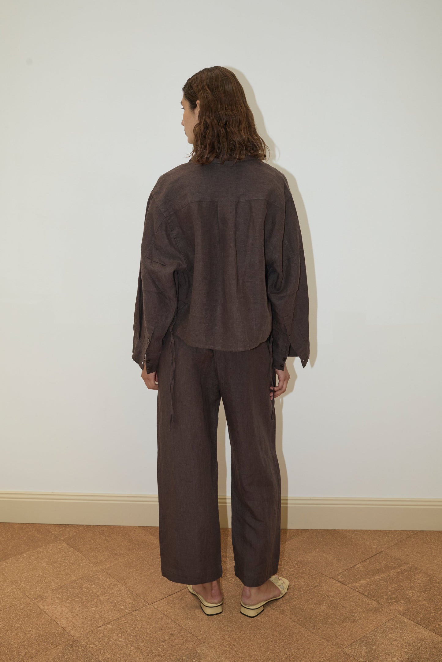 A person with wavy brown hair, wearing the Deiji Studios 4 Tie Set in Bark, stands facing away from the camera in a plain room with tan flooring and a white wall.
