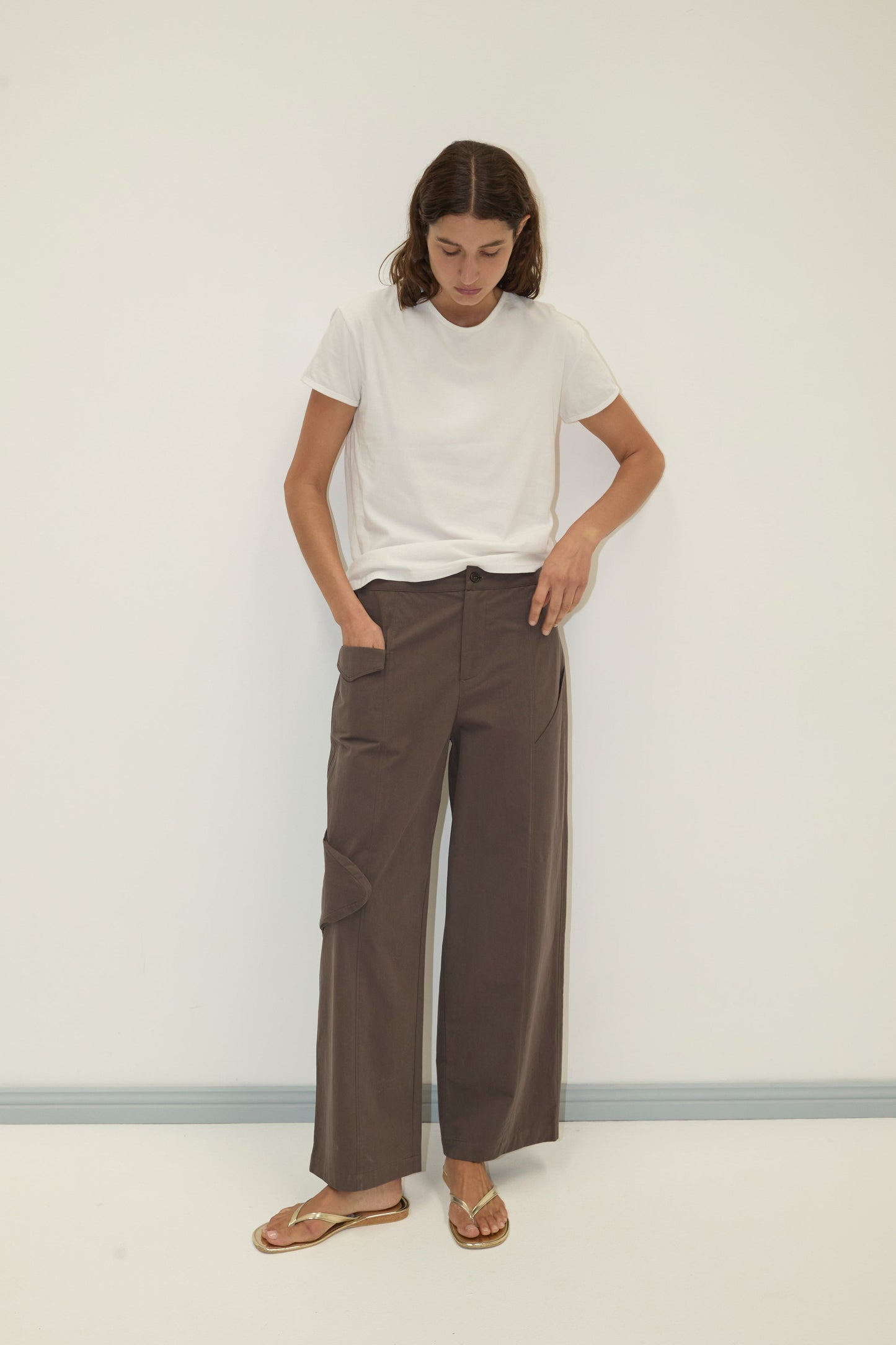 A person stands against a white wall, wearing a white t-shirt and the Pocket Pant - Wood: wide-leg brown organic cotton twill pants with side cargo pockets. Their hands are in their pockets and they are looking down.
