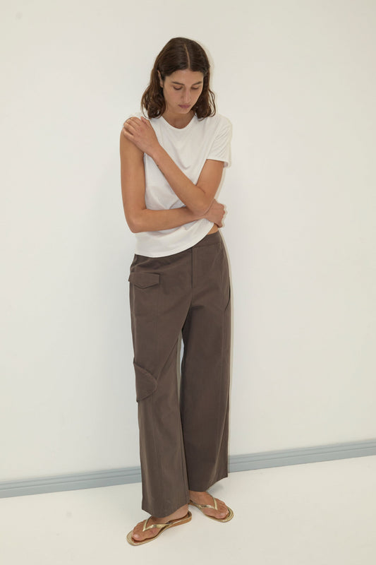 A person with long brown hair stands against a white wall, wearing a Deiji Studios white short-sleeve t-shirt, Pocket Pant - Wood, and tan sandals, with one arm crossed over their chest.