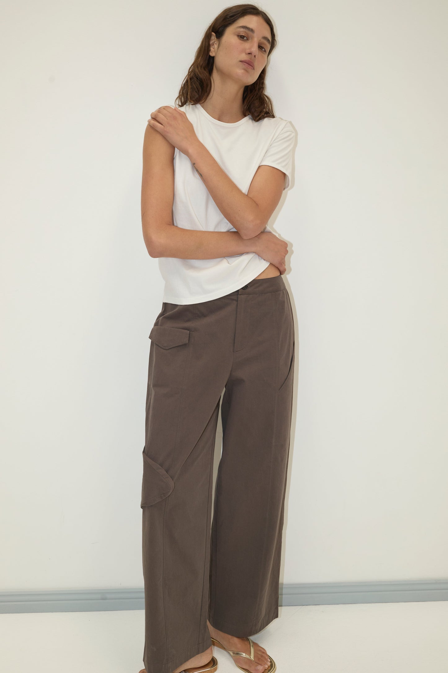 A woman with long brown hair stands against a white wall, arms crossed, wearing Deiji Studios' Simple Tee - White and loose brown pocketed pants, paired with gold sandals for a relaxed, signature look.