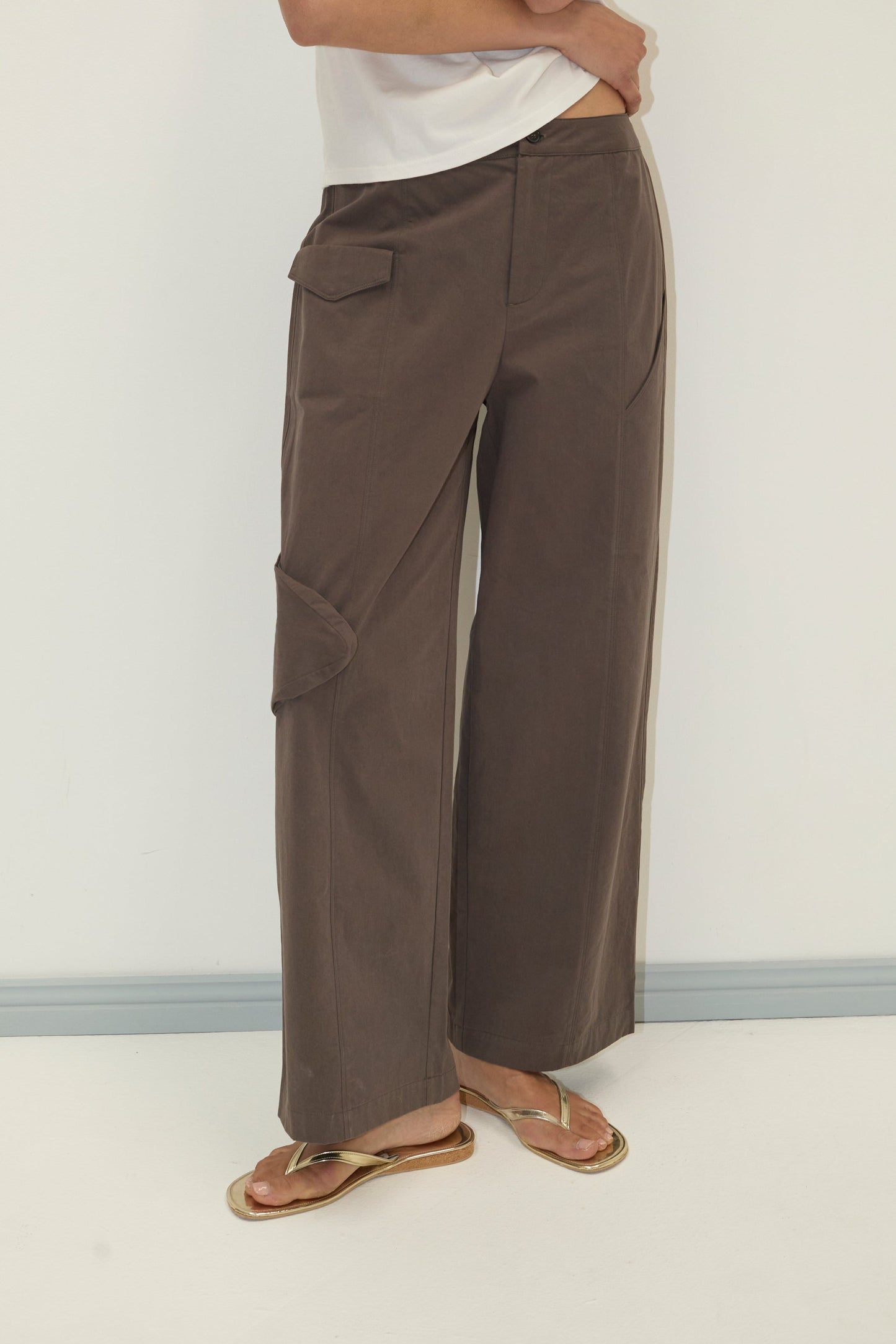 A person wearing the Pocket Pant - Wood, wide-leg brown cargo pants made from organic cotton twill, with a white top and gold sandals stands against a plain light wall. Their face is not visible.