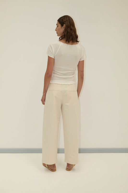 A person with shoulder-length brown hair stands facing away, wearing a short-sleeved white top, Flag Tie Pant in off white, and sandals against a plain light-colored background.
