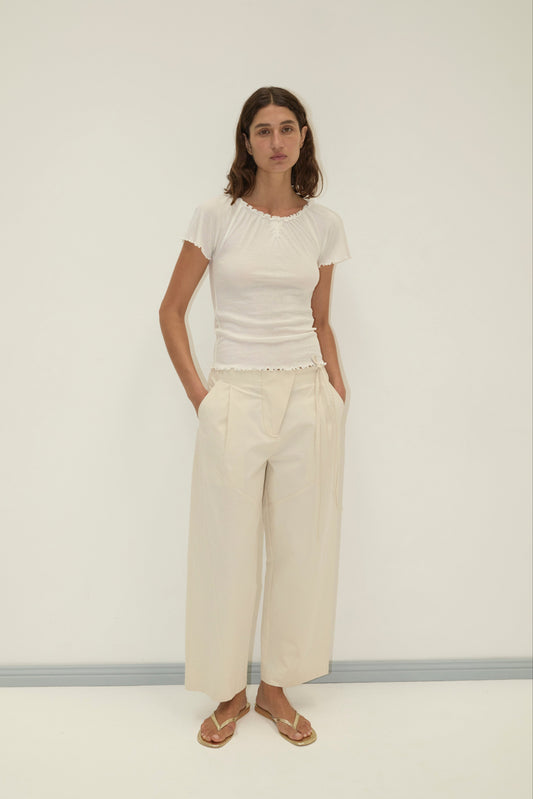 A woman stands against a white wall wearing a fitted white short-sleeve top, Flag Tie Pant in cream, and beige sandals. Her hands are in her pockets and she has a neutral expression.
