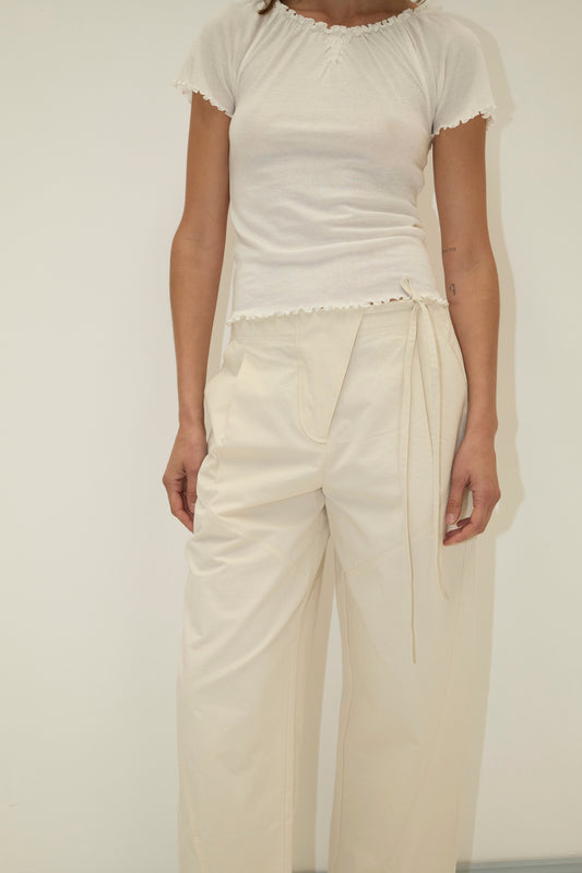 A person models the Flag Tie Pant - Off White, a pair of high-waisted, loose organic cotton pants, with a fitted white short-sleeve top against a plain light background. Their face is not visible.