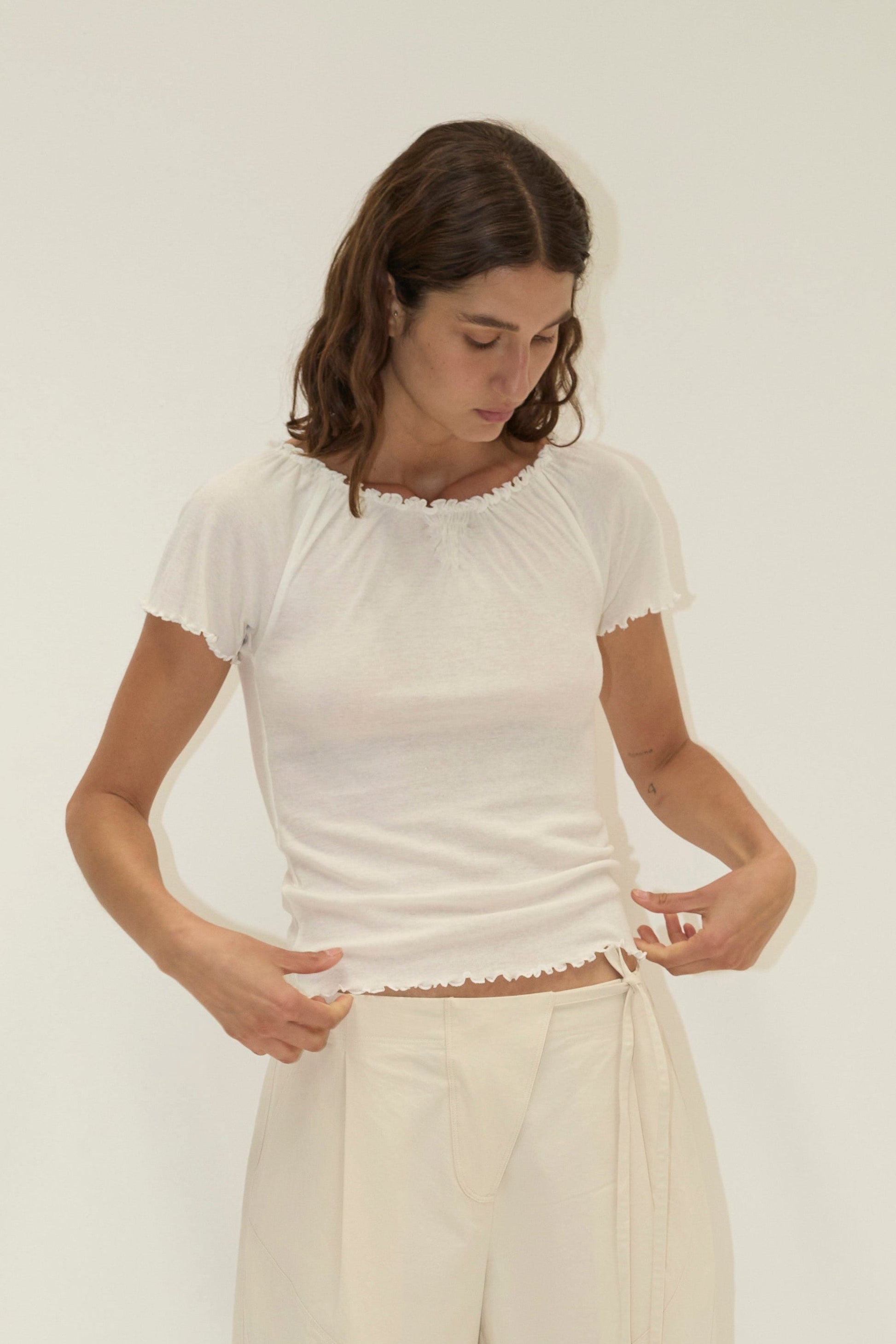 A woman with wavy brown hair wears the Flounce Tee - Cream by Deiji Studios, gently pulling on its hem as she looks down, standing before a plain off-white background.