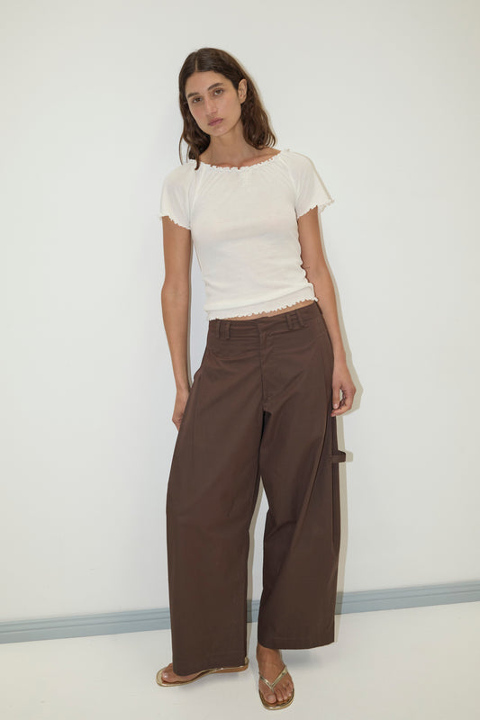 A woman with long brown hair and a neutral expression stands against a white wall, wearing a fitted white short-sleeve top, Deiji Studios Tab Pant in Dark Brown, and gold sandals.