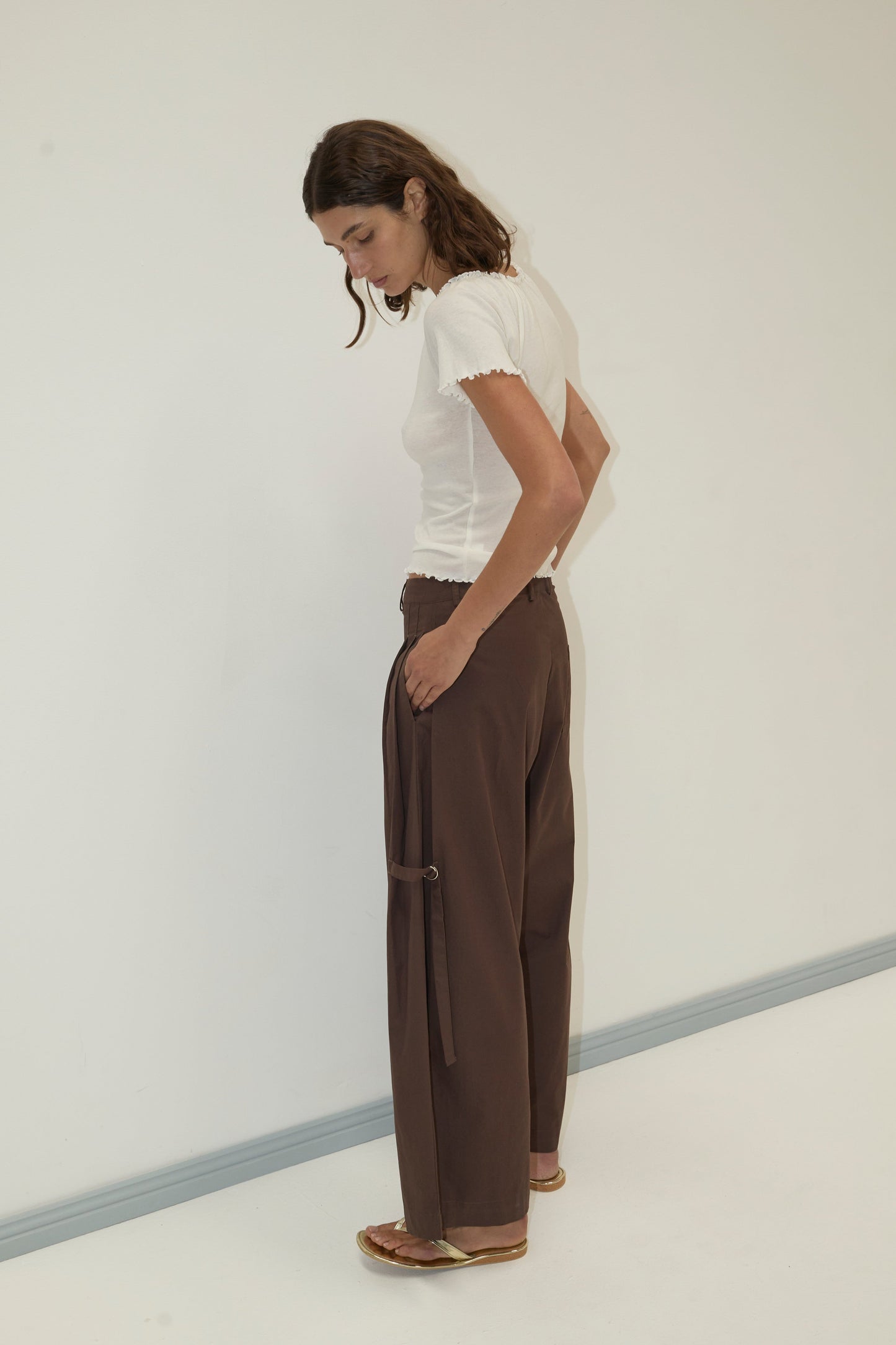 A woman with shoulder-length brown hair stands in a minimalist room, wearing a fitted white short-sleeve top and the Tab Pant - Dark Brown from Deiji Studios, featuring wide legs, side ties, and organic cotton. She looks down, body turned sideways.