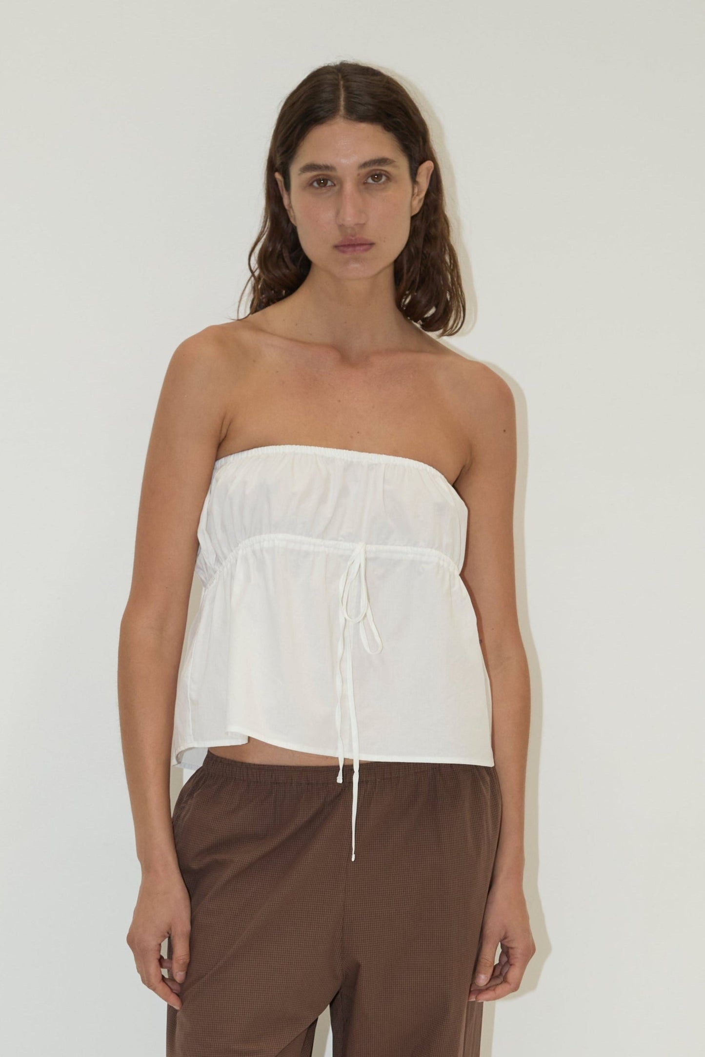A woman with long, wavy brown hair and a neutral expression wears a Deiji Studios strapless cotton top - white, crafted from organic cotton poplin with a drawstring waist, paired with loose brown pants against a plain white background.