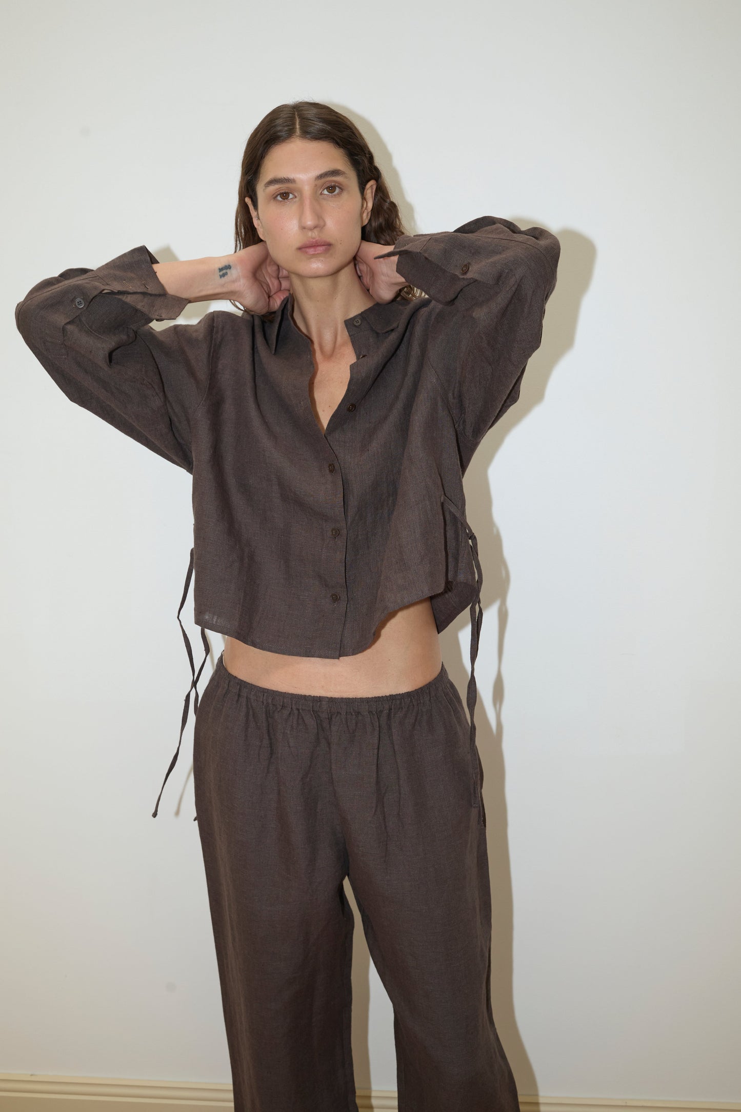A woman stands against a white wall wearing Deiji Studios' 4 Tie Set - Bark, a loose-fitting dark brown organic linen set featuring a long-sleeve button-up shirt and matching wide-leg pants, with her hands raised to touch her neck.