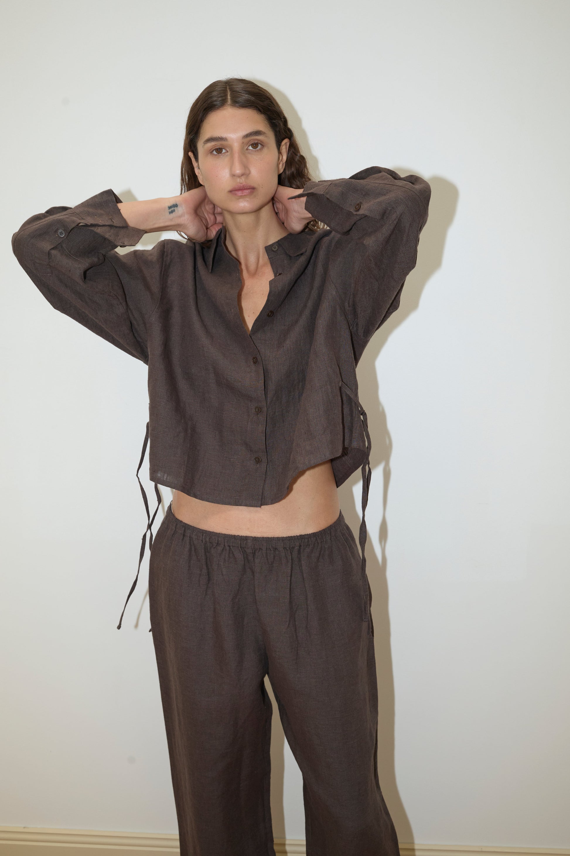 A woman stands against a white wall wearing Deiji Studios' 4 Tie Set - Bark, a loose-fitting dark brown organic linen set featuring a long-sleeve button-up shirt and matching wide-leg pants, with her hands raised to touch her neck.