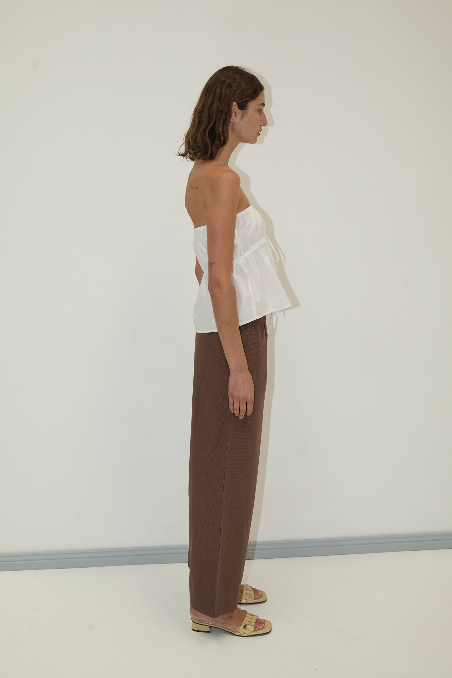 A woman with short brown hair stands in profile against a plain white wall, wearing a strapless white top, Deiji Studios Ease Trouser - Carob Check with an elastic waist, and beige slide sandals.