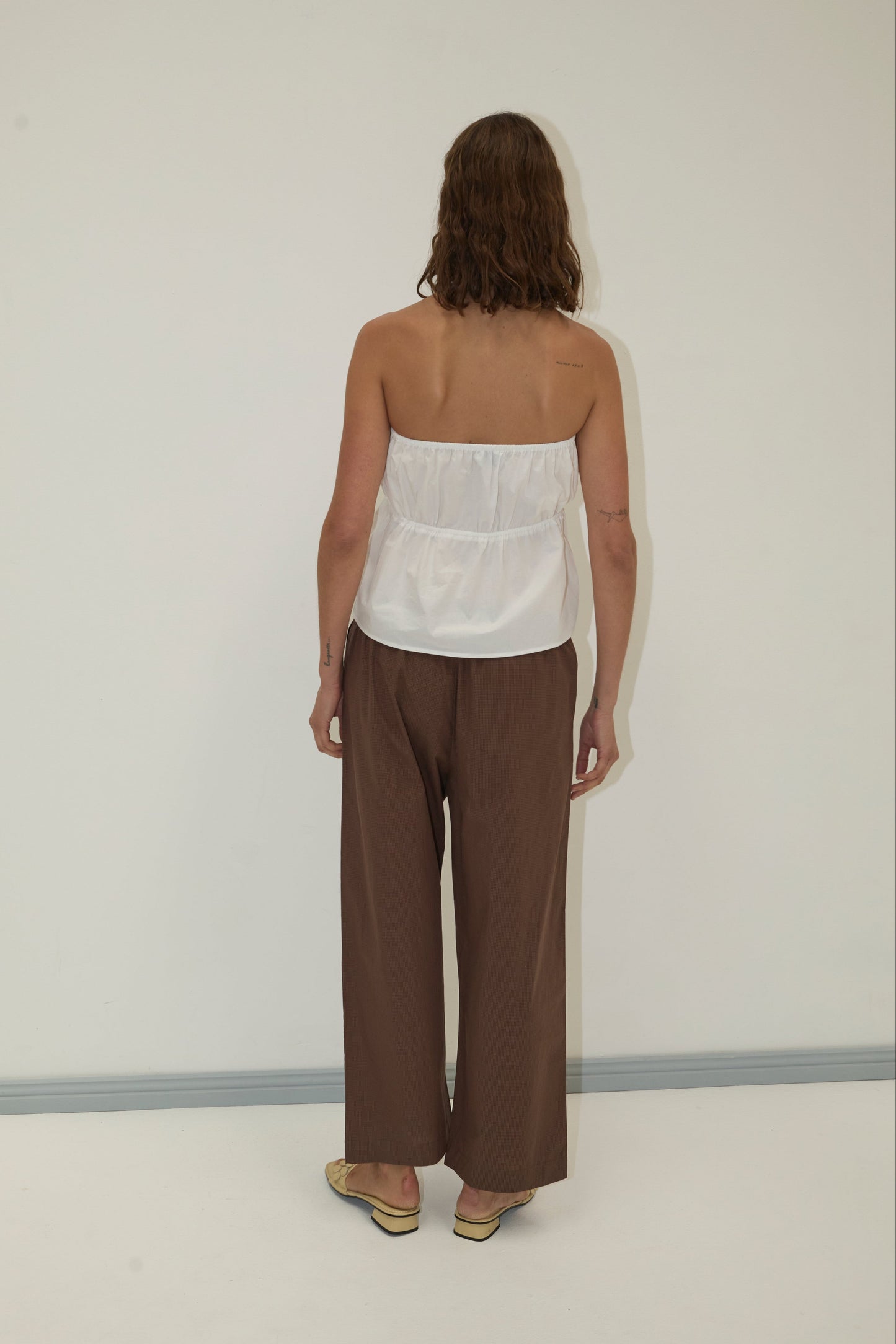 A person with wavy, shoulder-length hair stands facing a white wall, wearing a Deiji Studios strapless cotton top - white, loose brown organic cotton poplin pants, and beige shoes. Their arms are relaxed at their sides.