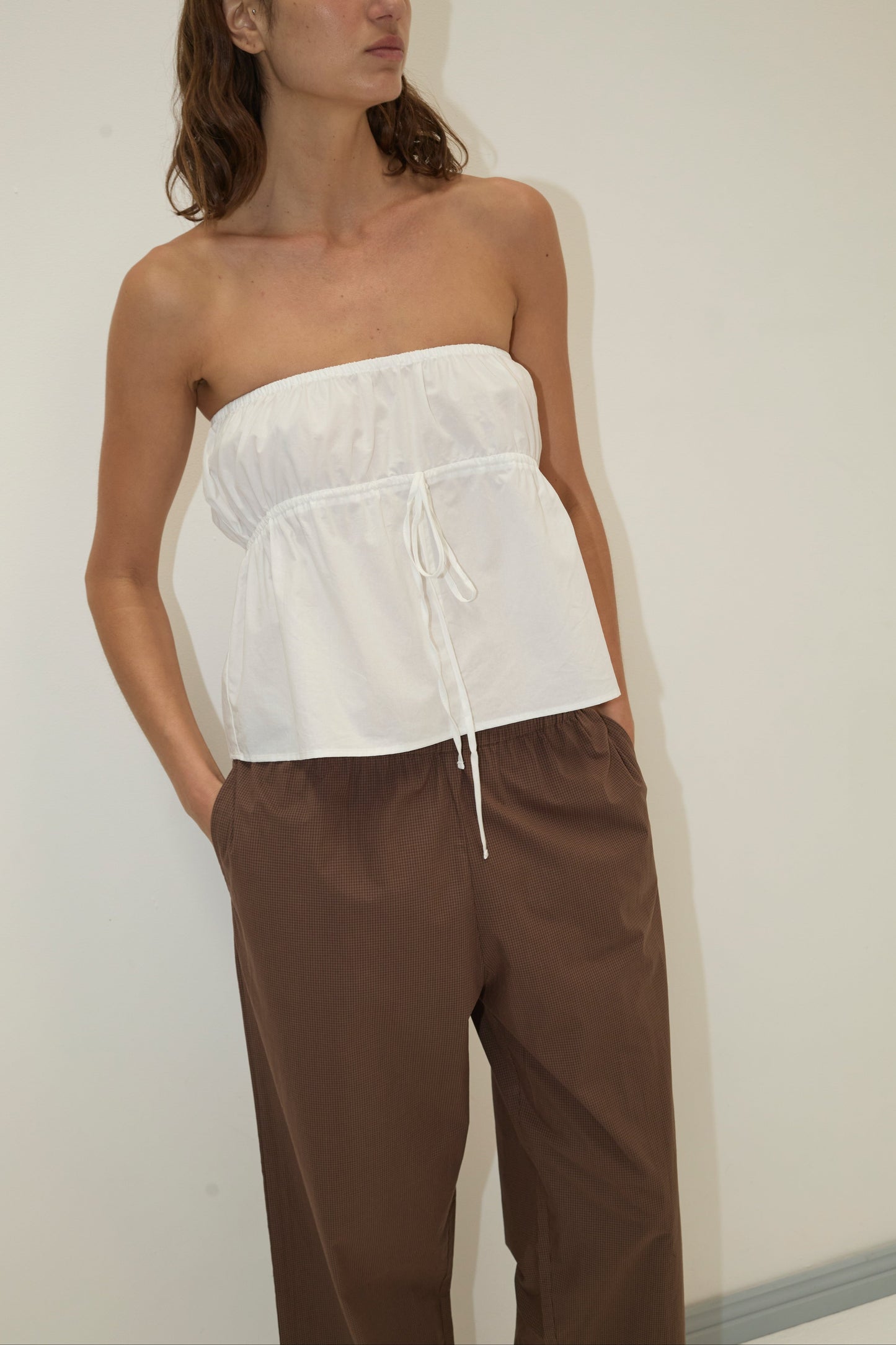 A person wearing the strapless cotton top - white stands against a plain light-colored wall, paired with loose brown pants, hands in pockets and head slightly turned to the side.