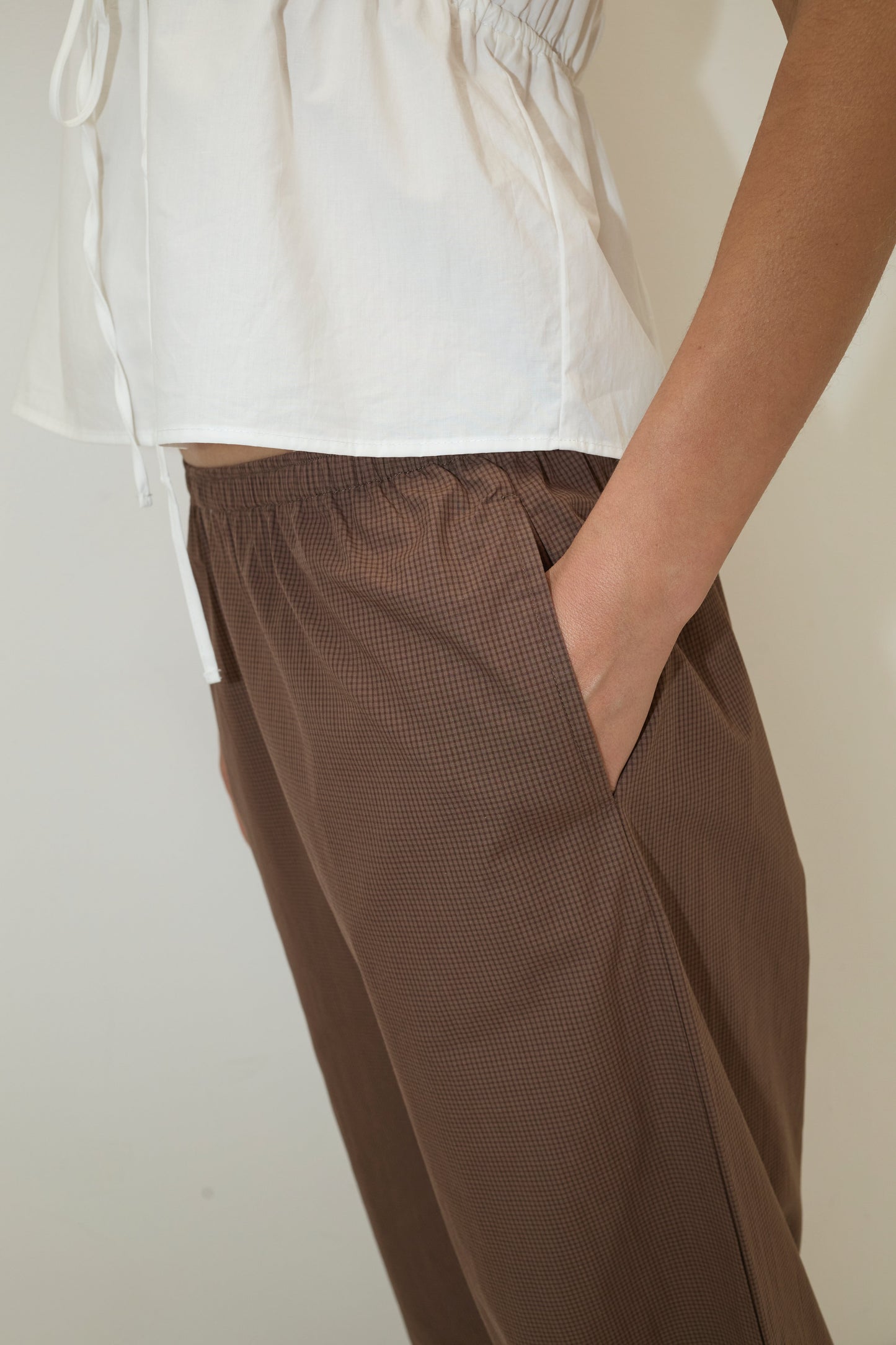 A person wears a white short-sleeved top and the Ease Trouser - Carob Check from Deiji Studios, standing with one hand in their pocket against a plain light backdrop. The image highlights the trouser’s relaxed fit through the torso and hips.