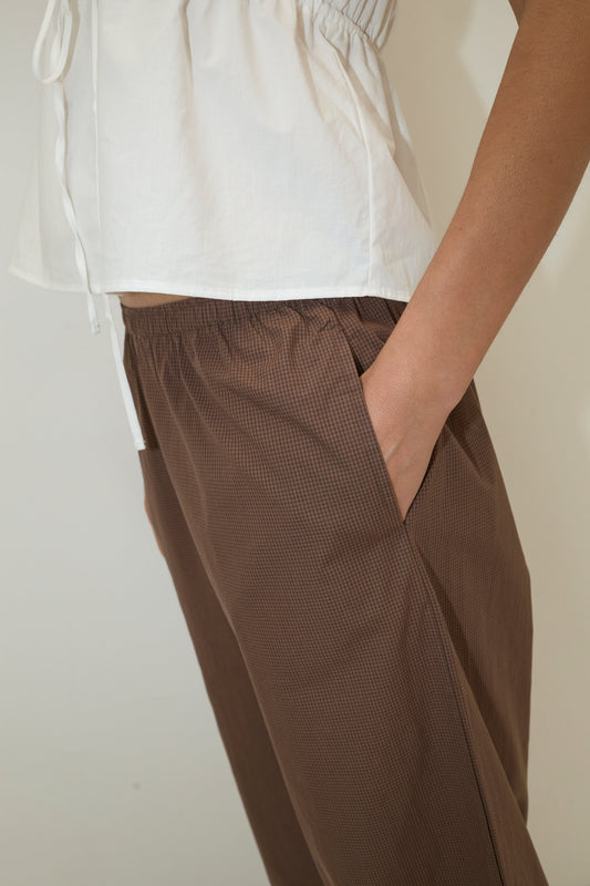 A person wears a white short-sleeved top and the Ease Trouser - Carob Check from Deiji Studios, standing with one hand in their pocket against a plain light backdrop. The image highlights the trouser’s relaxed fit through the torso and hips.