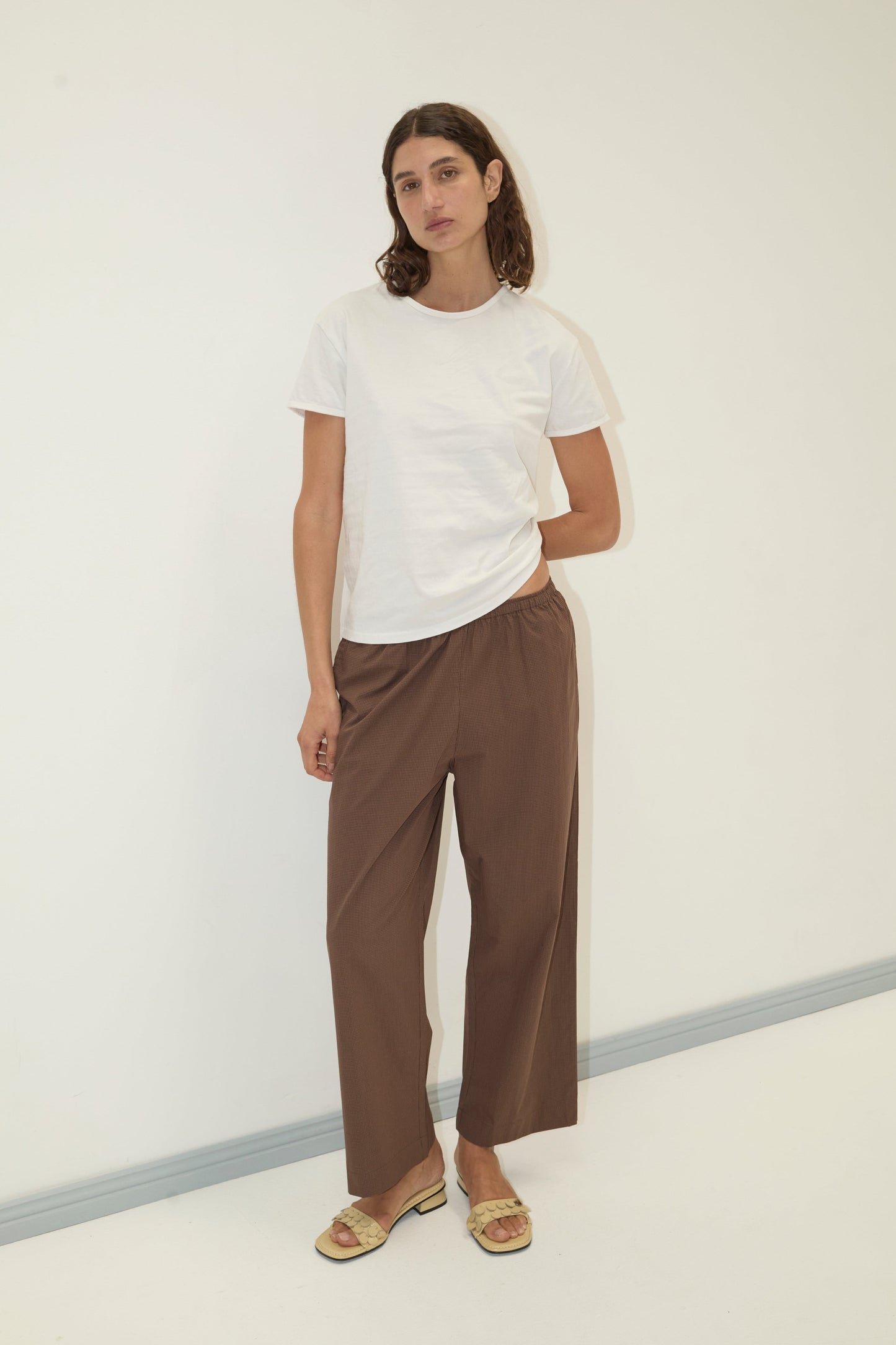 A person with shoulder-length brown hair and a neutral expression stands against a plain white wall, wearing the Ease Trouser in brown, a white t-shirt, and beige sandals.