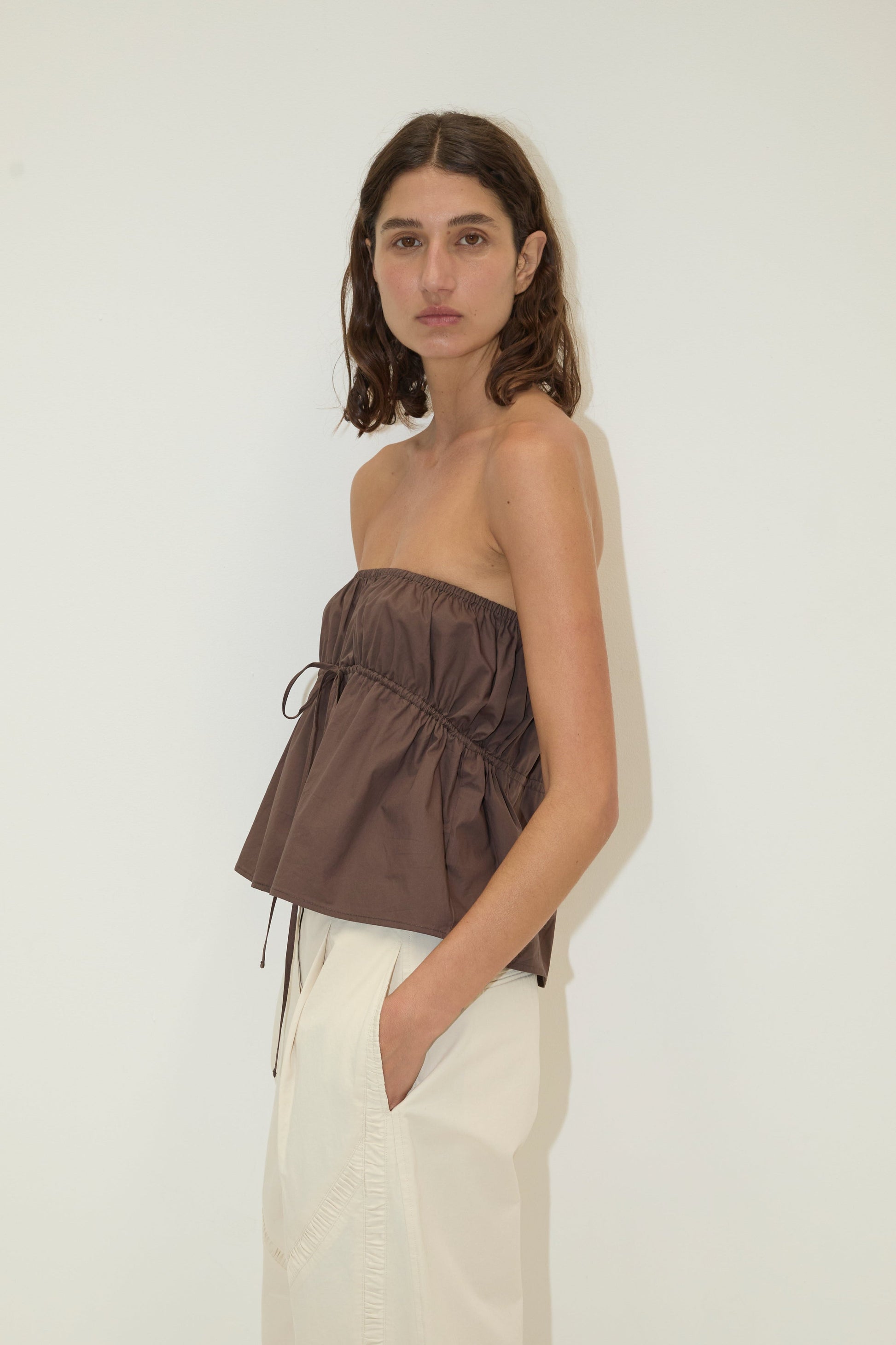 A woman with wavy brown hair stands against a white wall, wearing the Strapless Cotton Top in brown with a gathered waist and loose white pants, hands in her pockets, looking directly at the camera.