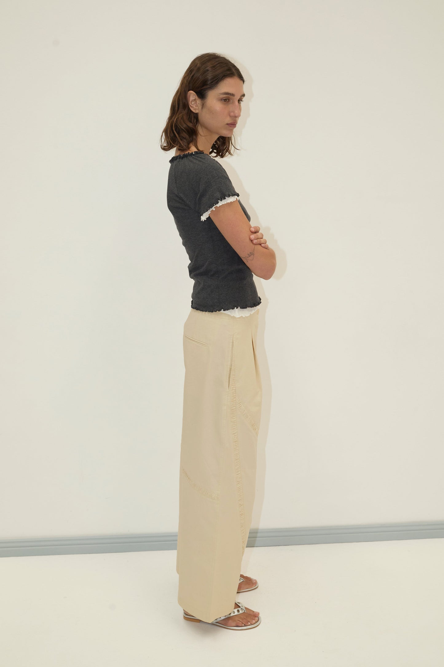 A woman with shoulder-length brown hair stands sideways against a plain white wall, arms crossed and neutral-faced. She wears a dark short-sleeve top, sandals, and the Channel Pant - Stone.