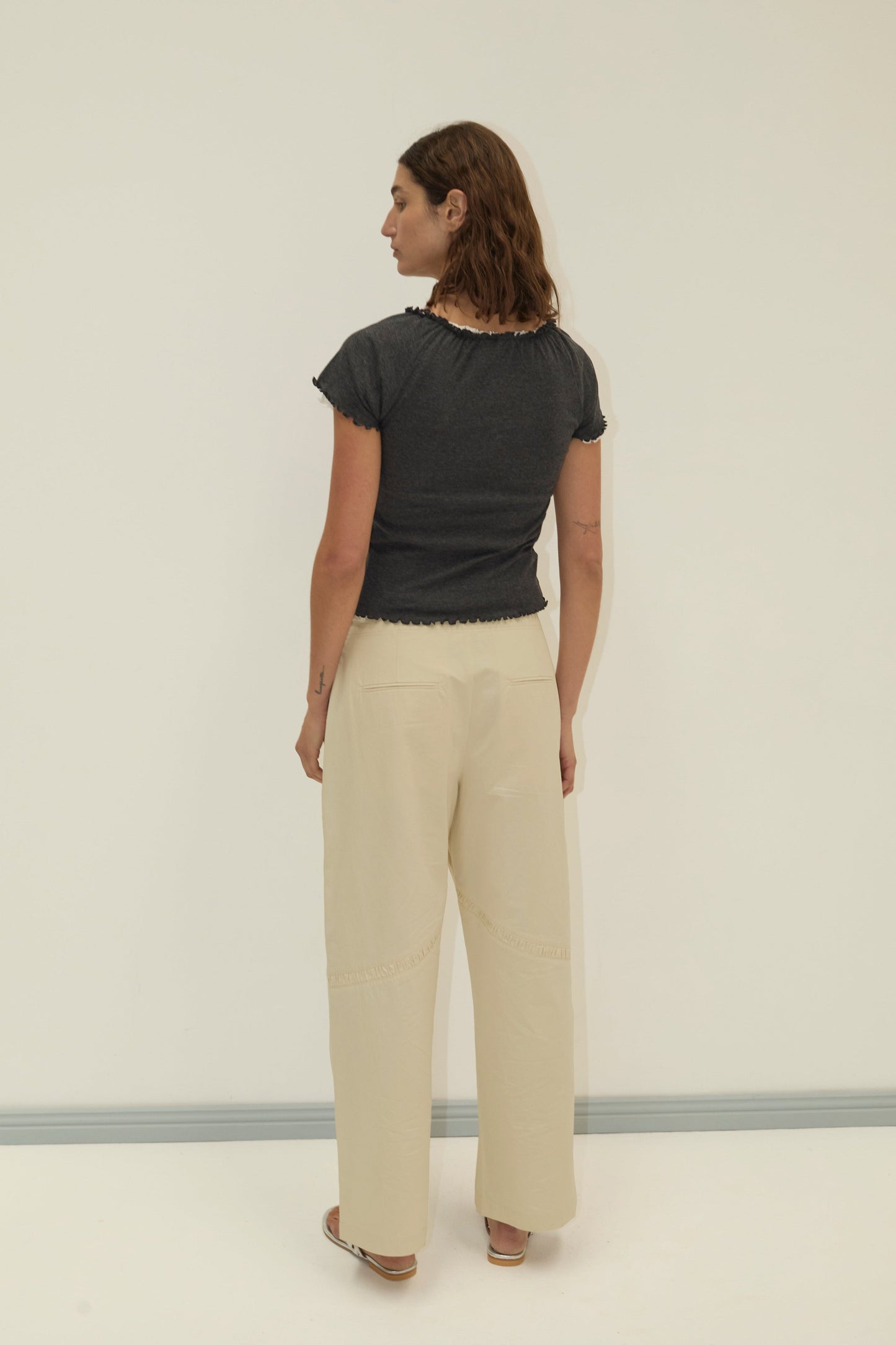 A person with shoulder-length brown hair stands facing away, wearing a fitted dark gray t-shirt, Channel Pant - Stone by Deiji Studios in beige, and brown sandals against a plain light background.