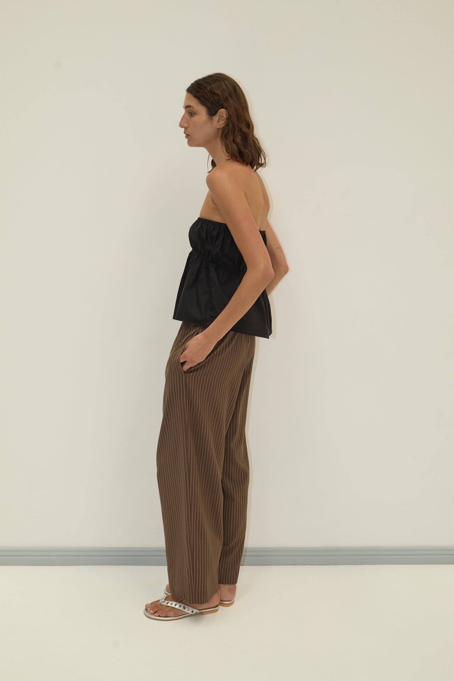 A person stands in profile against a plain light wall, wearing a black strapless top, Pure Wool Ease Trouser - Sable Stripe by Deiji Studios, and flat sandals. Their brown hair is shoulder-length.