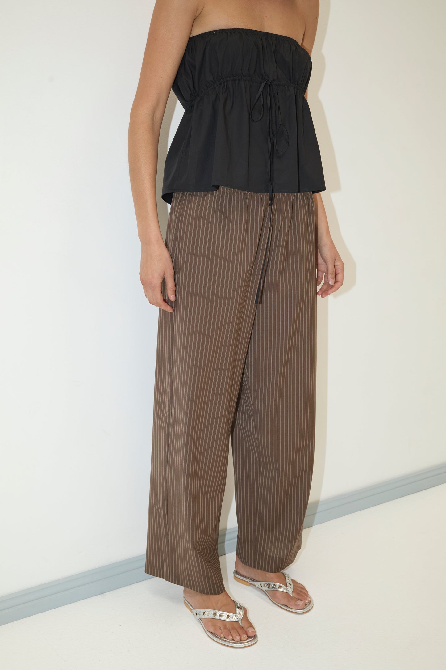 A person wearing a black strapless top, Pure Wool Ease Trouser - Sable Stripe from Deiji Studios, and white sandals stands against a plain white wall with arms relaxed by their sides.