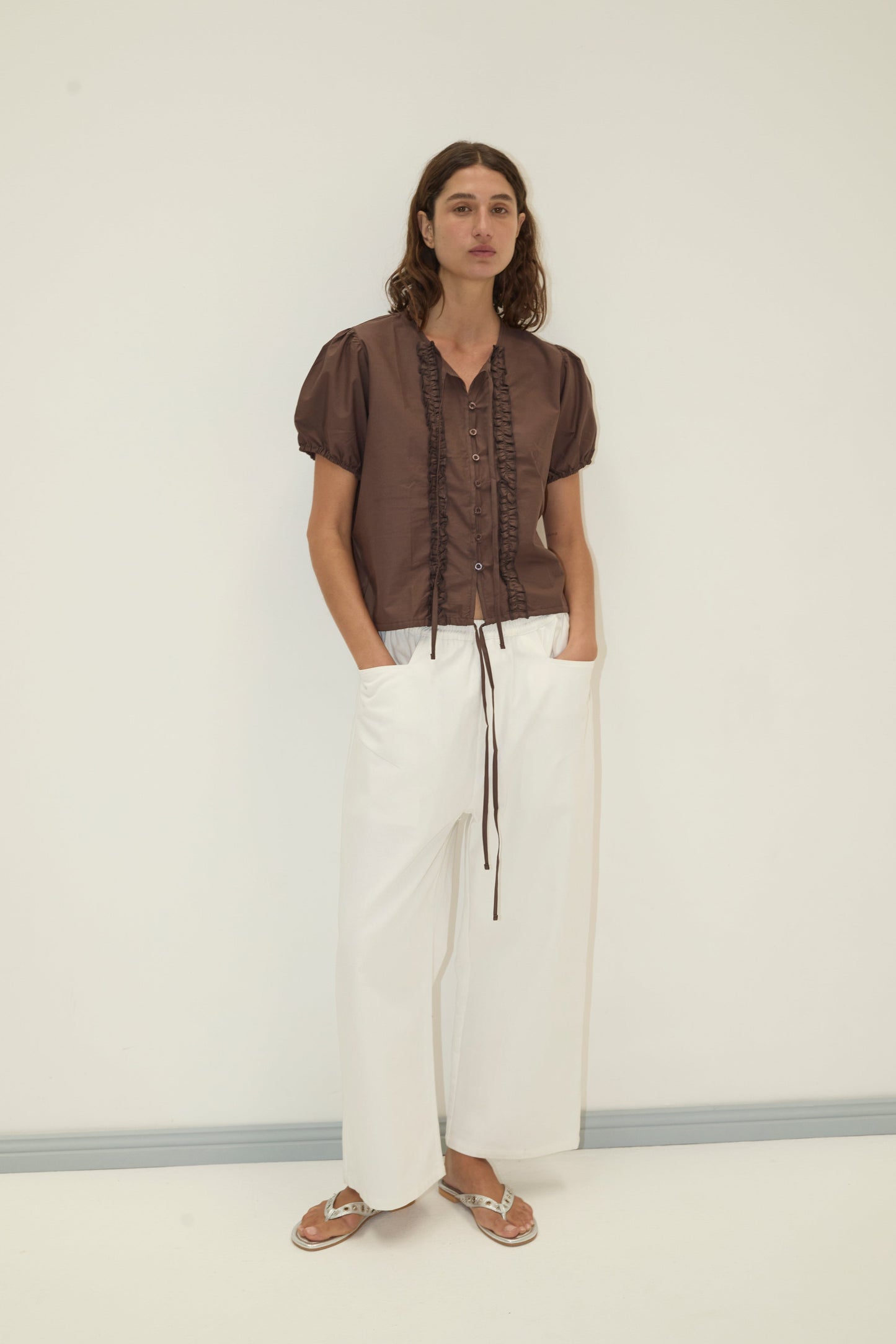 A person stands against a plain wall wearing the Double Frill Top - Chocolate by Deiji Studios, paired with loose white pants and silver sandals. Their hands are in their pockets and they have long, wavy hair.