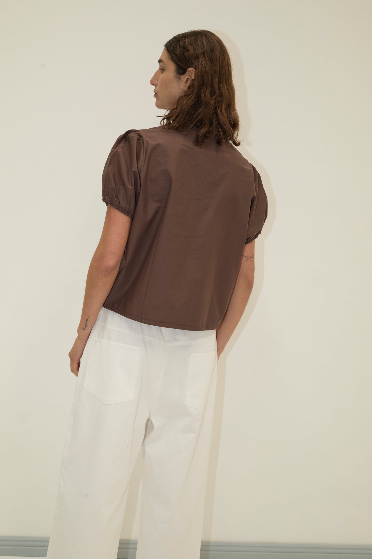 A person with shoulder-length brown hair stands facing away, wearing the Double Frill Top - Chocolate by Deiji Studios in organic cotton and high-waisted white wide-leg pants against a plain light background.