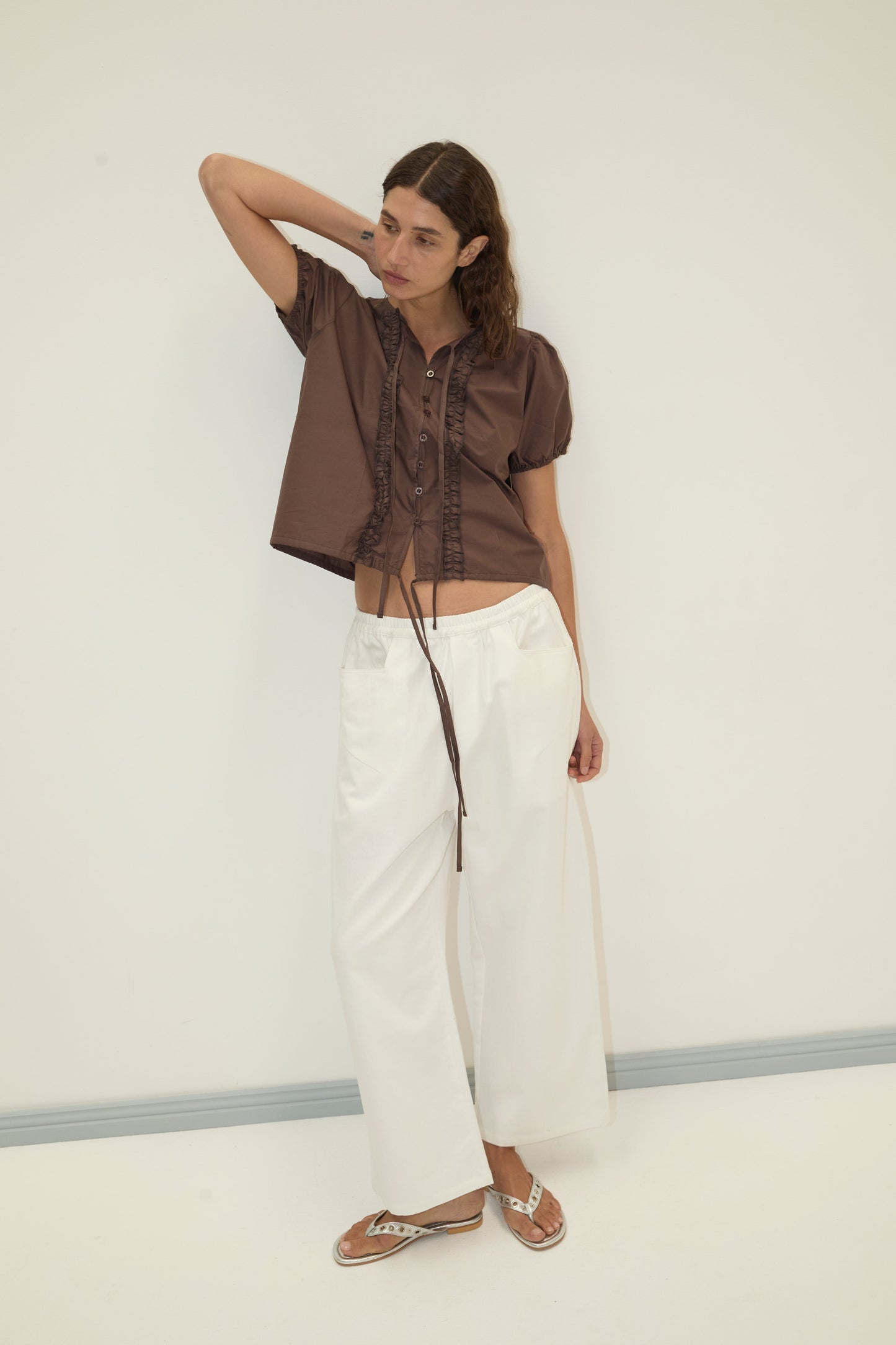 A woman stands against a white wall, wearing the Double Frill Top - Chocolate by Deiji Studios with tie details, paired with loose white pants and sandals. She rests one hand behind her head and looks to the side.