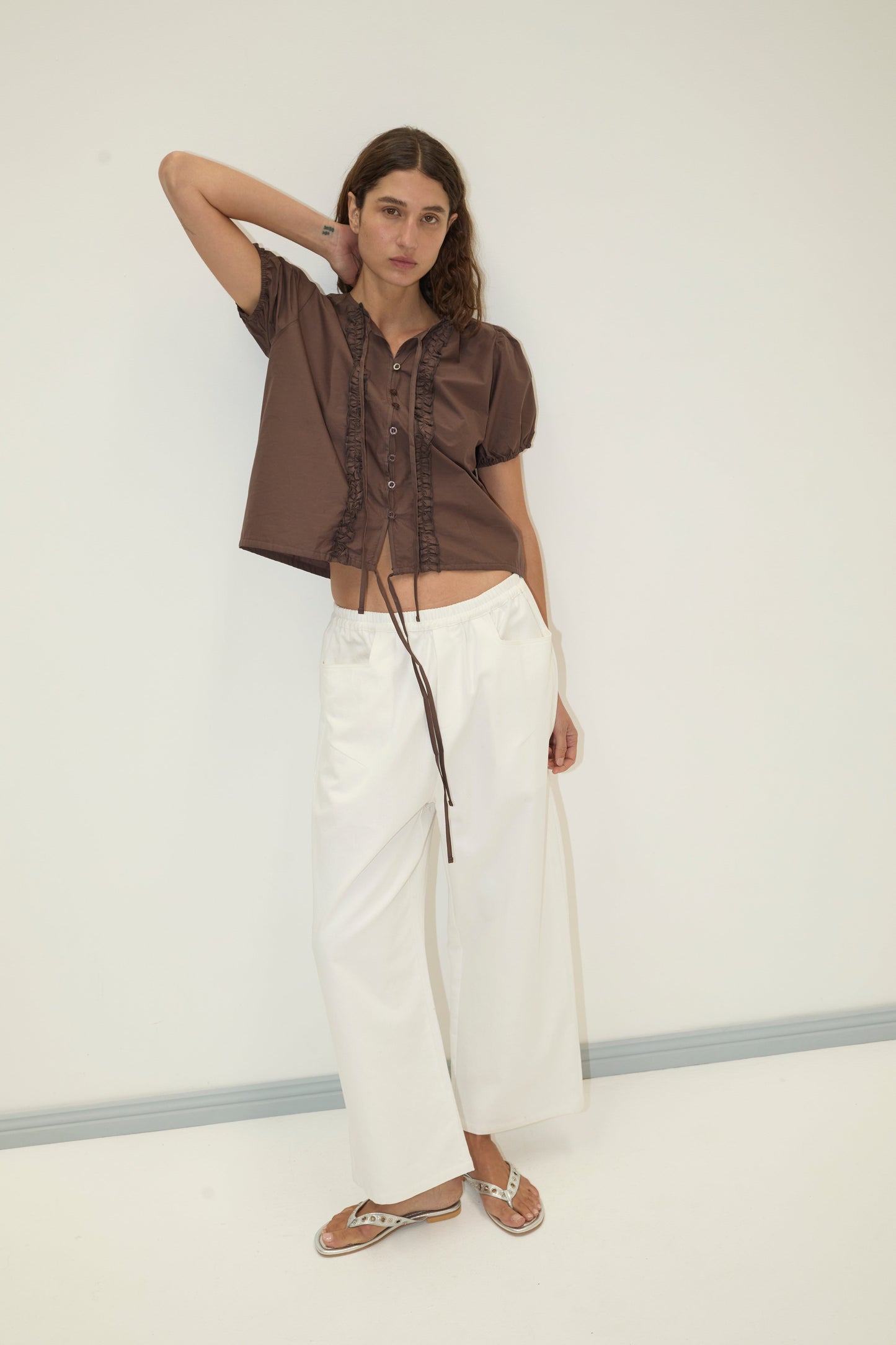 A woman stands against a white wall wearing the Square Pocket Pant in white, a brown short-sleeve blouse, and flip-flops. She has one hand behind her head and maintains a neutral expression.