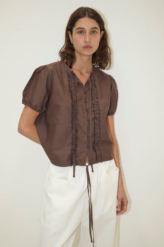 A person with wavy brown hair wears the Double Frill Top, a brown short-sleeve blouse with ruffled trim, and white pants while standing against a plain light background.