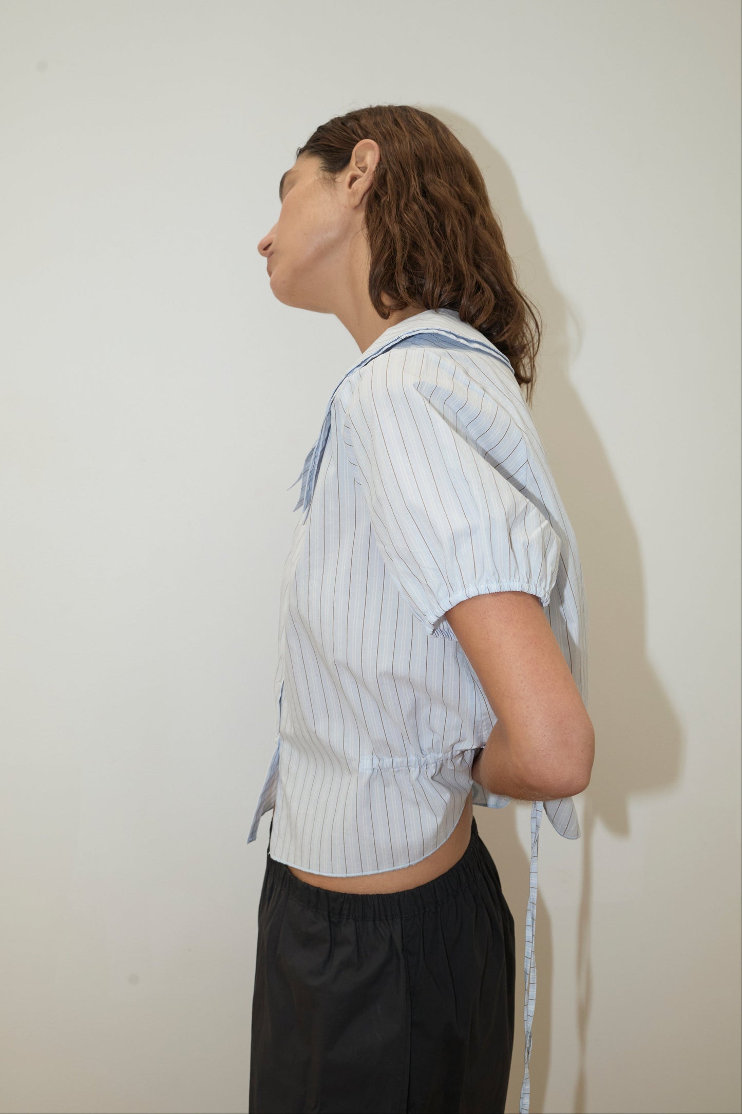 A person with wavy brown hair wears Deiji Studios' Double Collar Shirt - Mineral Stripe, light blue and short-sleeved, paired with dark pants. They stand sideways against a plain light wall, one arm tucked behind their back.