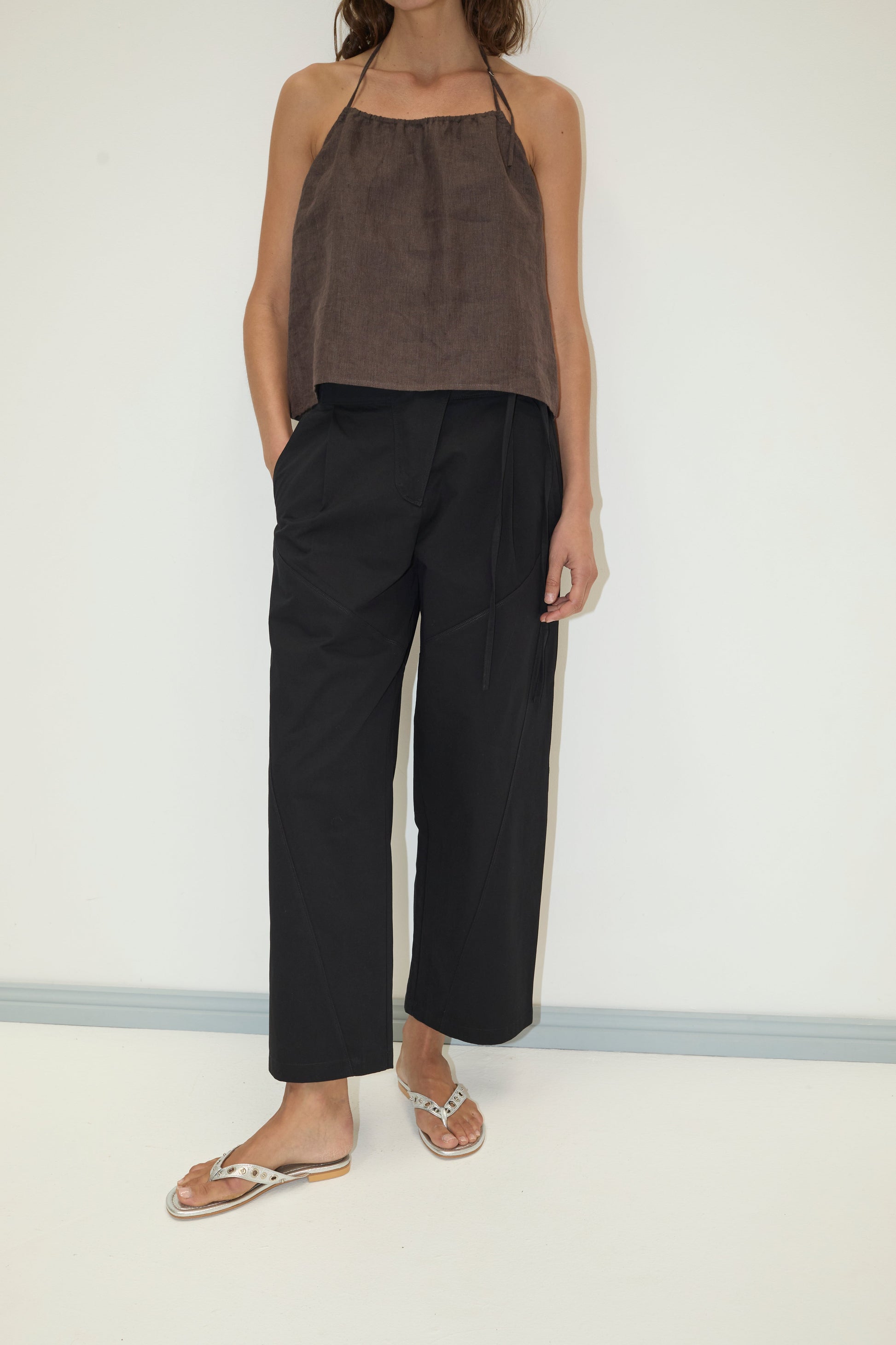 A woman stands against a white wall wearing black Flag Tie Pant with pockets, a brown halter top, and white sandals. Her right hand is in her pocket; her head is partially out of frame.