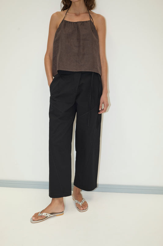 A woman stands against a white wall wearing black Flag Tie Pant with pockets, a brown halter top, and white sandals. Her right hand is in her pocket; her head is partially out of frame.