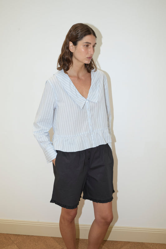Model wearing Flounce Short - Black with lace trim and relaxed fit, paired with a light blue pinstripe long-sleeve V-neck top with ruffled hem, standing against a white wall.