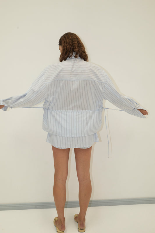 Back view of a model in the Deiji Studios Room Shirt - Mineral Stripe, an oversized light blue pinstripe long sleeve shirt jacket with tie detail and matching mini skirt, gold mule heels, arms outstretched against a cream wall.