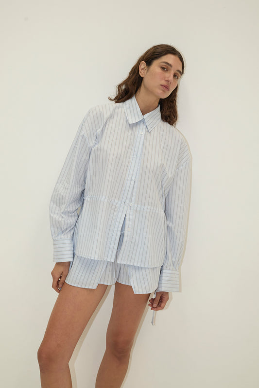 A model wears Deiji Studios' Room Shirt - Mineral Stripe, a light blue and white pinstripe oversized long-sleeve collared shirt with matching mini shorts, styled against a white background.