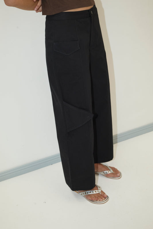A person wears the Pocket Pant - Black, featuring wide-leg organic cotton twill and angled pockets, paired with a brown top and rhinestone-detailed silver flip-flops, standing by a white wall with a gray baseboard.