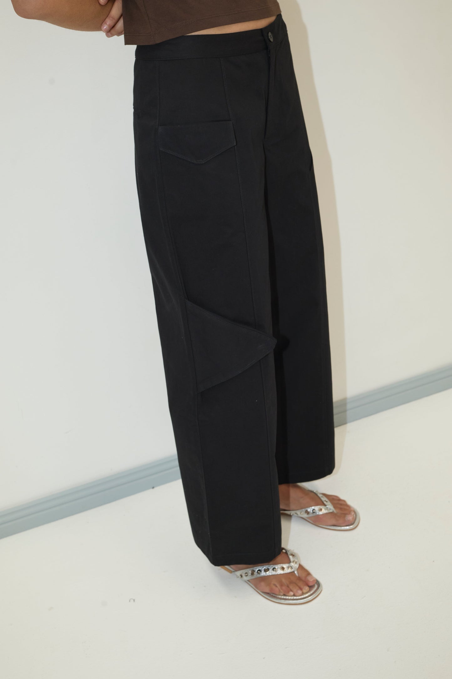 A person wearing the Pocket Pant - Black by Deiji Studios, a brown top, and white flip-flops with silver details stands on a white floor next to a gray baseboard. The image is cropped at the torso.
