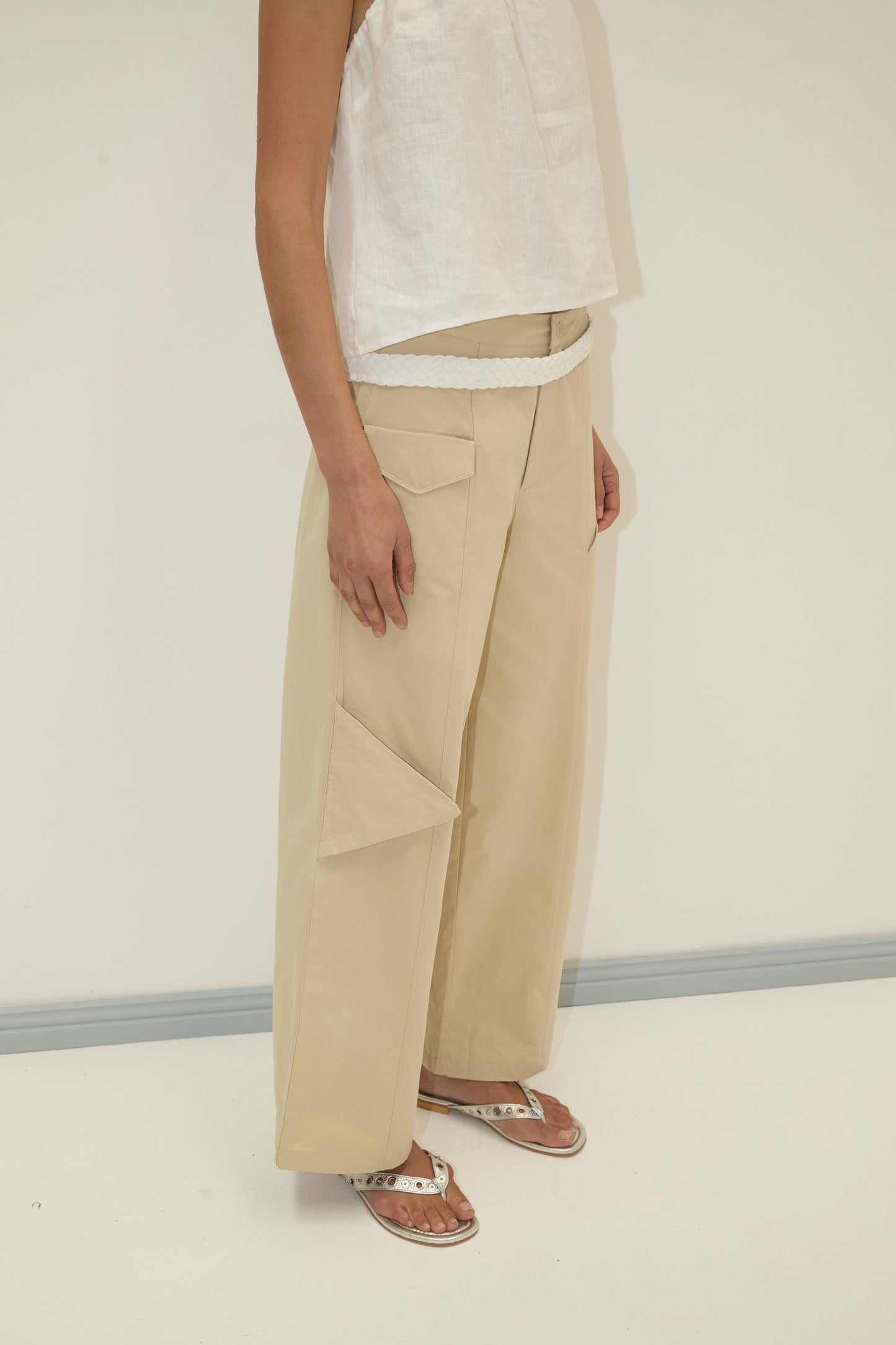A person wearing a sleeveless white top, Deiji Studios Pocket Pant - Stone with a large side pocket, a white braided belt, and white sandals stands against a plain light background.