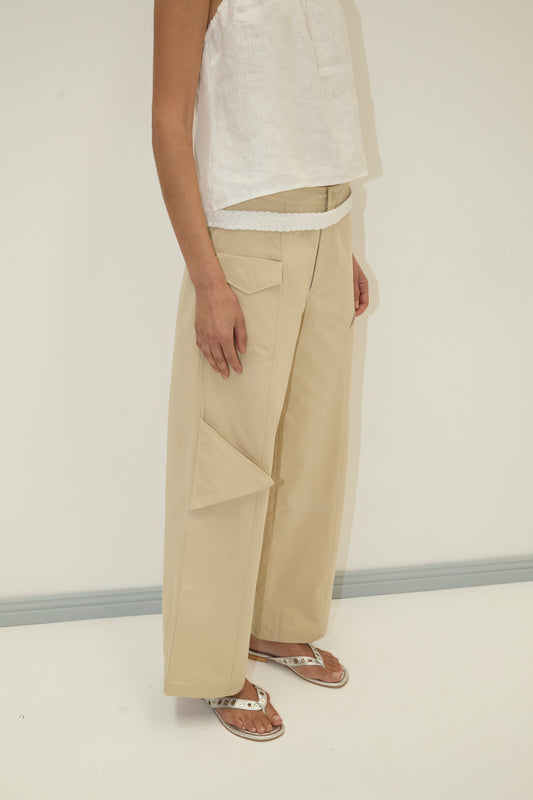 A person wearing a sleeveless white top, Deiji Studios Pocket Pant - Stone in organic cotton twill with a side pocket, a white belt, and white sandals stands against a plain white background.