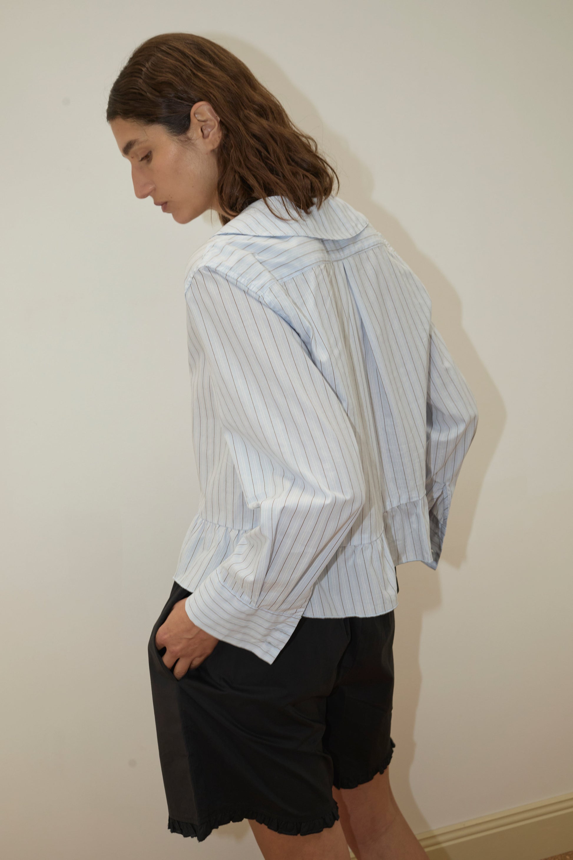 Model seen from behind in an oversized white pinstripe shirt with a ruffled hem, paired with Flounce Short - Black featuring an elastic waist and flounced hem, hand in pocket, looking down.